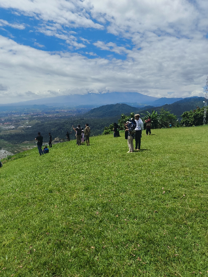 A group of people stand on a grassy hilltop overlooking a valley and distant mountains. There are around 12 people, some are standing and talking, others are looking out at the view, and a few are taking photos. There are trees and shrubs in the background and a clear blue sky with white clouds. The location is a hilltop in Majalengka, Indonesia, at coordinates -6.846, 108.204. The view from the hilltop is expansive, with the city of Majalengka stretching out in the distance, and mountains beyond. There is a transmission tower on the right side of the image in the distance, which is a common sight in rural Indonesia. The scene appears peaceful and serene, with the people enjoying the natural beauty of the surrounding landscape.