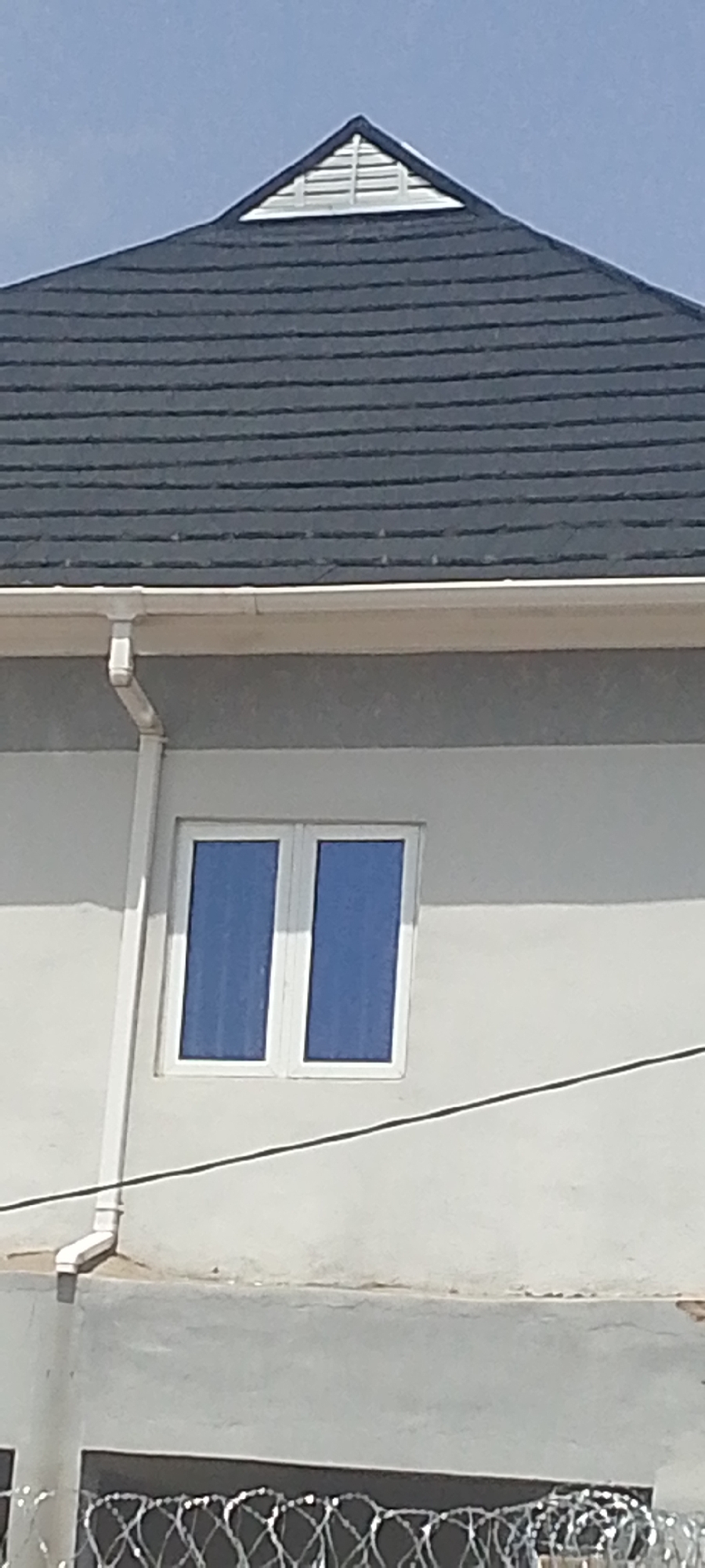 The image shows the upper part of a building's exterior on a bright, sunny day. A dark grey, shingled roof slopes upwards to a triangular gable vent, which is white and louvered. Below the roofline, a beige gutter runs horizontally, and a white downspout descends vertically along the pale grey wall. A double-paned window with blue-tinted glass is centered on the wall. The lower portion of the image reveals a barbed wire fence. The scene suggests an architectural or construction context.