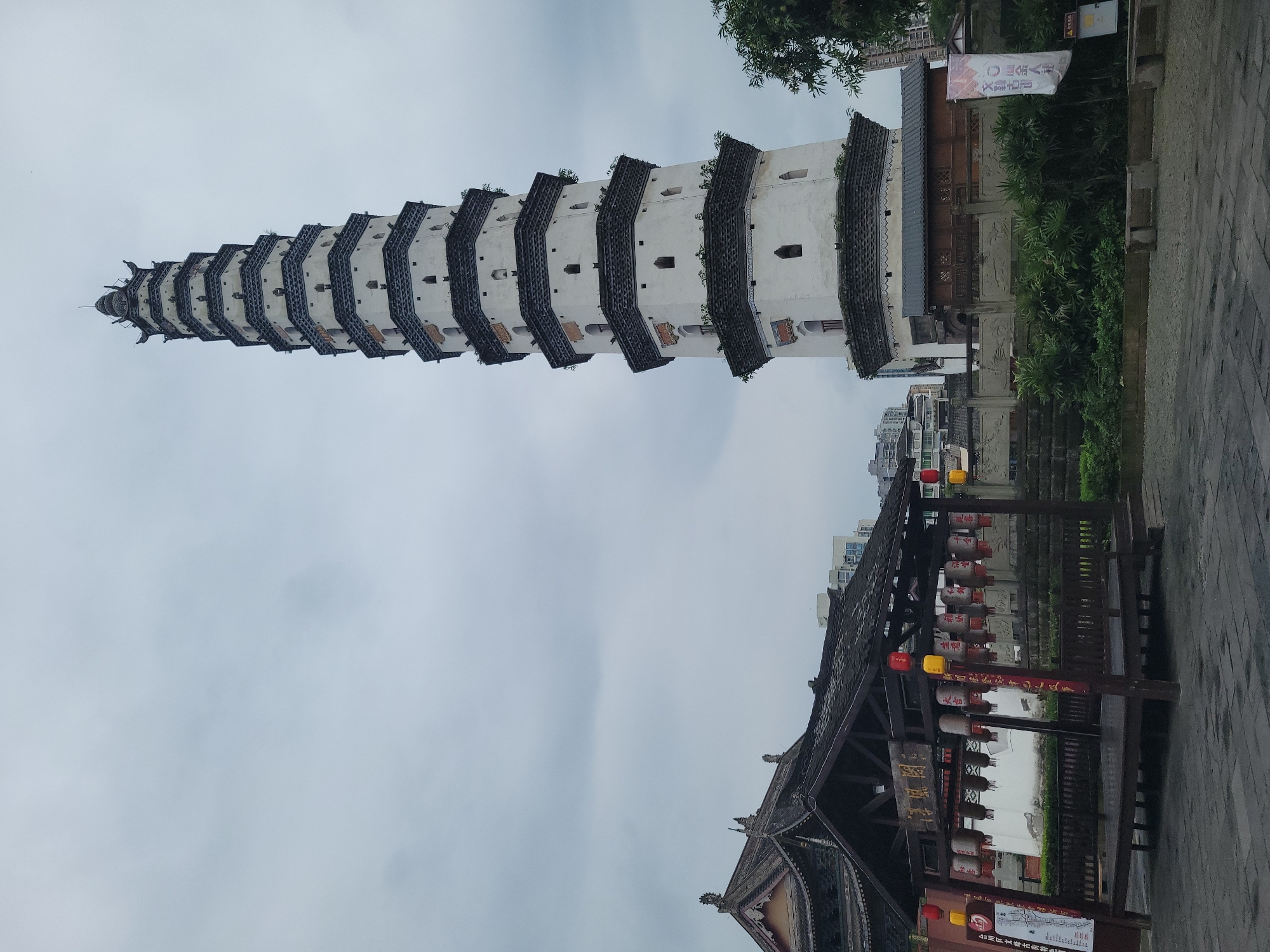 Here's a description of the image:
The image shows a tall, multi-tiered pagoda-style tower in Hechuan, China. The tower is white with dark, protruding roof sections on each level. It extends vertically into a gray, overcast sky. At the base of the tower is a traditional Chinese structure with lanterns and signage in front of it, indicating a commercial area in the ancient town of Wen, Hechuan. The environment is partly urban, with buildings visible in the background. The image appears to be a street-level view.