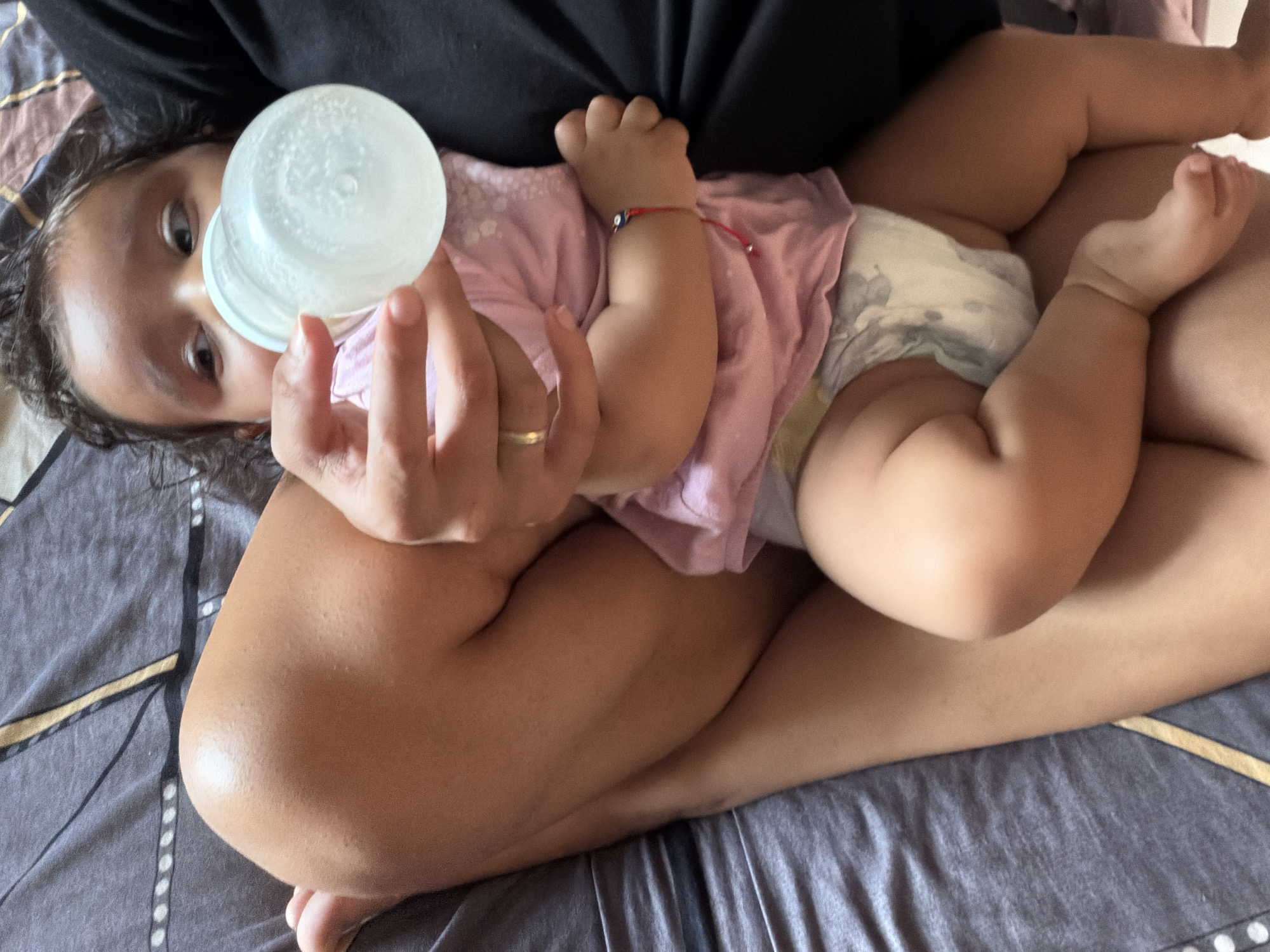 A close-up, overhead shot shows a baby being fed from a bottle while held on someone's lap. The baby is wearing a pink shirt and a diaper. The person holding the baby is wearing a black shirt. The background is a bed with gray and gold patterned sheets. The location is Razhevo Konare, Bulgaria.