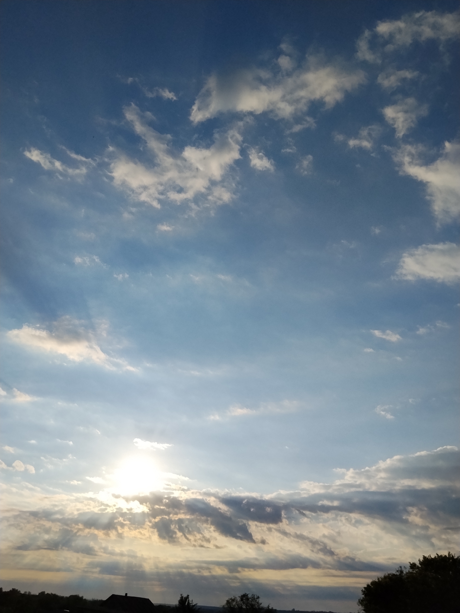 The image captures a sky filled with scattered clouds at sunset over Ustyvytsia, Ukraine. The sun is partially obscured by clouds, casting rays of light and creating a dramatic effect. Silhouetted against the sky are trees and a building with a dark roof, indicating a rural or semi-rural setting. The overall scene is tranquil, with the vibrant colors of the sunset and the interplay of light and shadow.