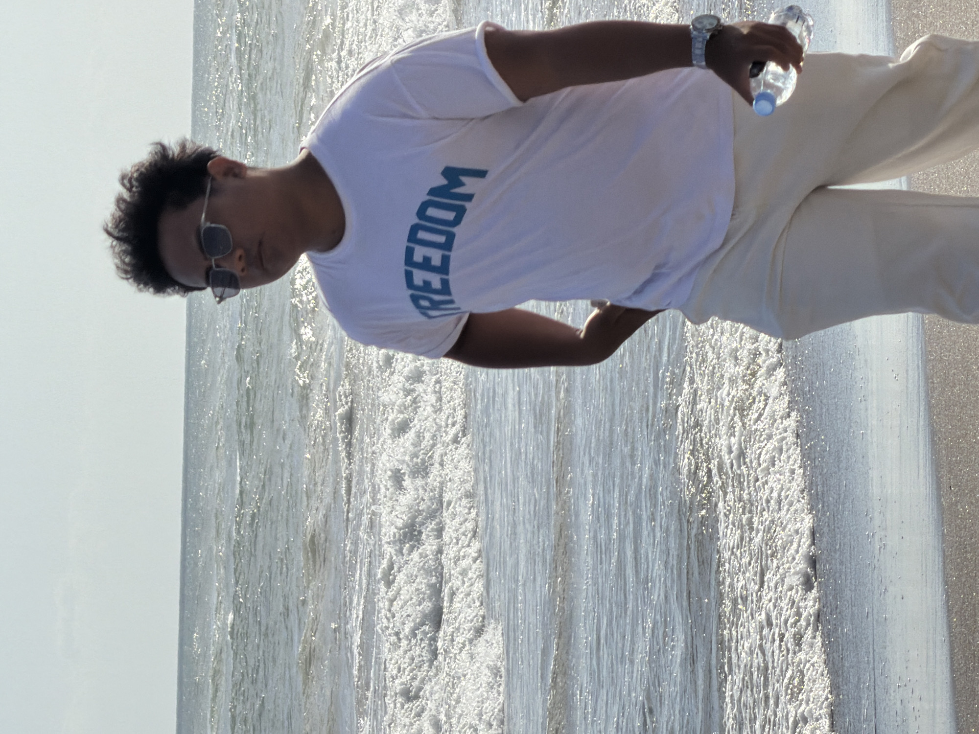 A person walks along the beach in North Kuta, Indonesia. The person is wearing a white t-shirt with the word "FREEDOM" printed in blue on the front, sunglasses, and light-colored pants. They are holding a clear plastic water bottle in their right hand and have their left hand in their pocket. The ocean with moderate waves is visible behind them, with sunlight glinting off the water. The sky is clear and light. The sand on the beach is dark.