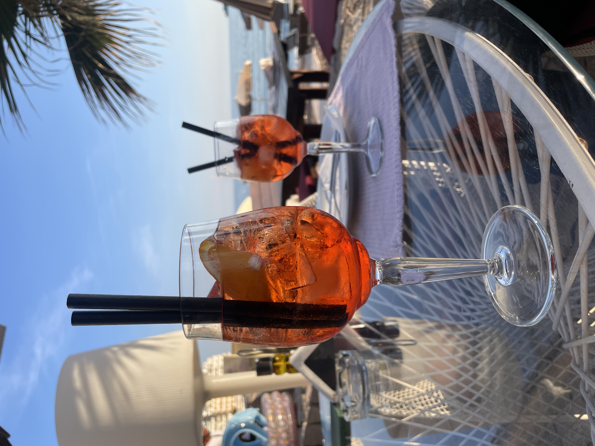 The image displays an outdoor scene, likely at a coastal bar or restaurant in Budva, Montenegro, under a clear blue sky. Two stemmed glasses, each containing a vibrant orange-red cocktail, are prominently featured on a glass-top table. The cocktails, resembling Aperol Spritz, are filled with ice cubes and garnished with a slice of citrus, probably orange, and each has two black straws.
The table beneath the drinks has a reflective glass surface and a distinctive white, woven base visible through the glass. A light lavender napkin is placed beneath one of the cocktail glasses. In the upper left quadrant, the fronds of a palm tree are silhouetted against the bright, expansive blue sky. In the background, beyond the table, a large body of water—presumably the sea—is visible, confirming a waterfront or beachside setting. Blurred elements further in the background suggest additional seating or structures of an outdoor venue. The scene conveys a relaxed, leisurely atmosphere on a sunny day. There is no visible text.