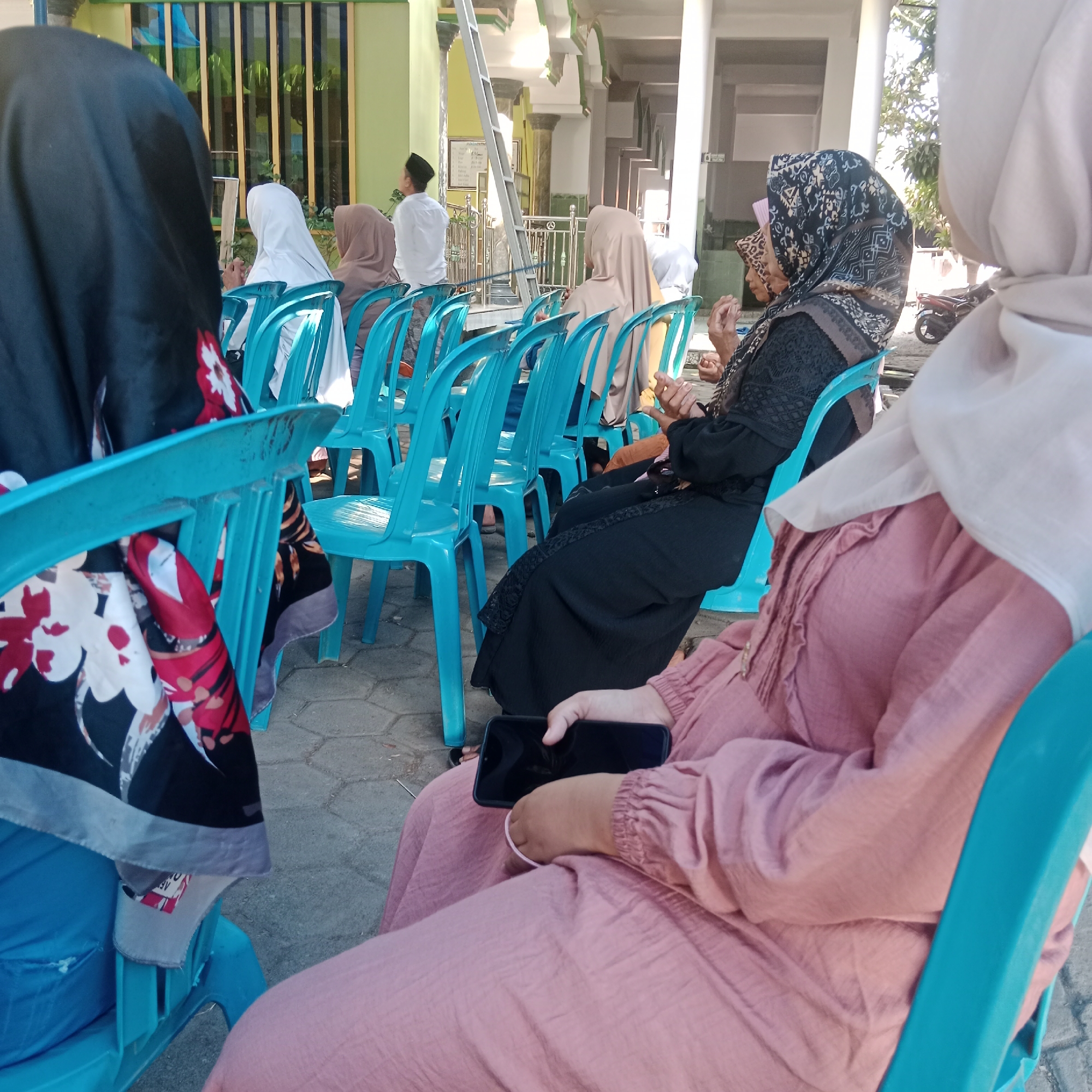 In Tegowanu Kulon, Indonesia, this image captures an outdoor gathering of people, primarily women, seated on turquoise plastic chairs arranged in rows. The setting appears to be in front of a building with light green and white architectural elements, suggesting it might be a place of worship or a community center. The ground is paved with light-colored rectangular stones. The individuals are dressed in Islamic attire, with many wearing hijabs of various colors and patterns, including black, white, beige, and patterned designs. Some women are engaged in conversation, while others are looking forward or down at their phones. One woman in the foreground is holding a black smartphone. In the background, a man in a white shirt and black cap is visible, along with a motorcycle parked to the right. The overall atmosphere suggests a community event or religious gathering taking place during daylight.