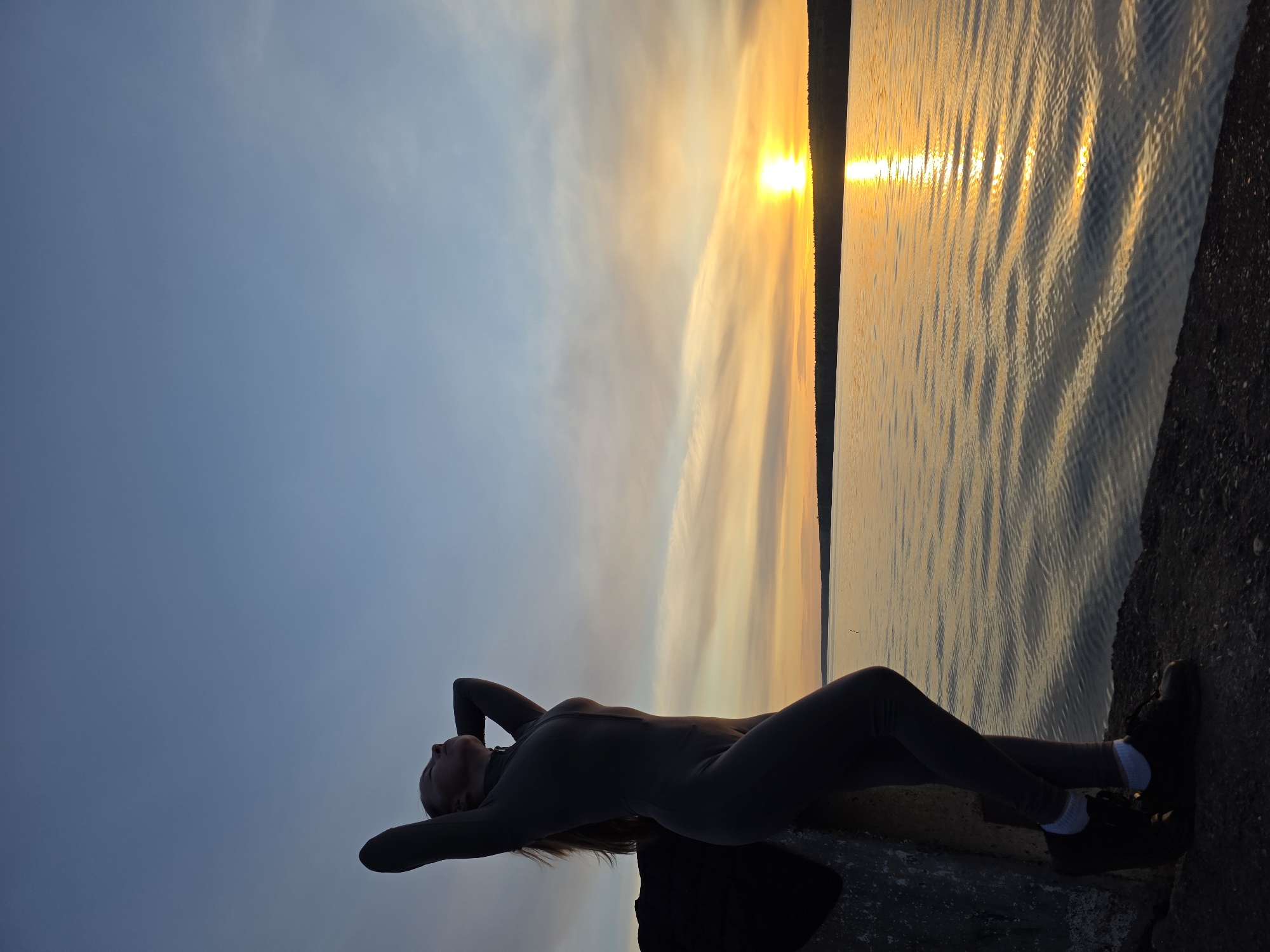 A woman is lying on a concrete structure next to a body of water at sunset. Her body is silhouetted against the colorful sky, with her head tilted back and her hands behind her head. She is wearing a full-body suit and sneakers with white socks. The sun is setting over the water, casting a bright reflection. Based on the surrounding environment (a large body of water and a cityscape in the distance), this image might have been taken at the Kama reservoir, a popular destination for locals and tourists.