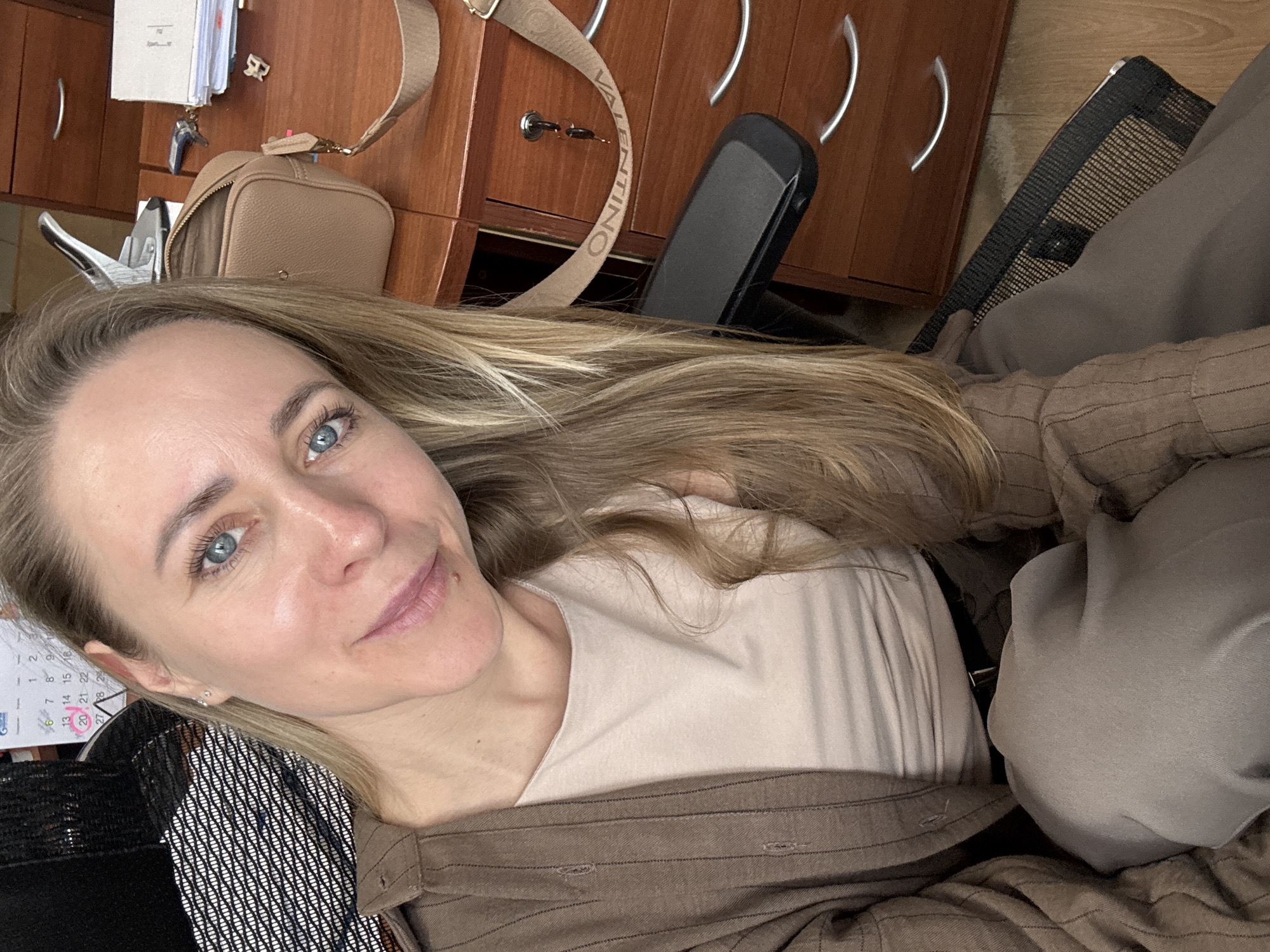 A woman with blonde hair and blue eyes is positioned in a high-angle selfie. She wears a light beige shirt under a brown striped button-down shirt, and sits in what appears to be an office chair.
Behind her is a desk with wooden cabinets. Items such as a small beige purse with a "VALENTINO" strap, office supplies like a stapler and documents, and keys are visible on the desk.
A portion of a calendar is also visible, showing some Russian script and a marked date of the 20th.
While it is not possible to confirm with certainty the exact location within the city of Chaikovsky, Russia based on the visual information alone, the calendar, office setting, and the woman's general appearance are the primary elements.
