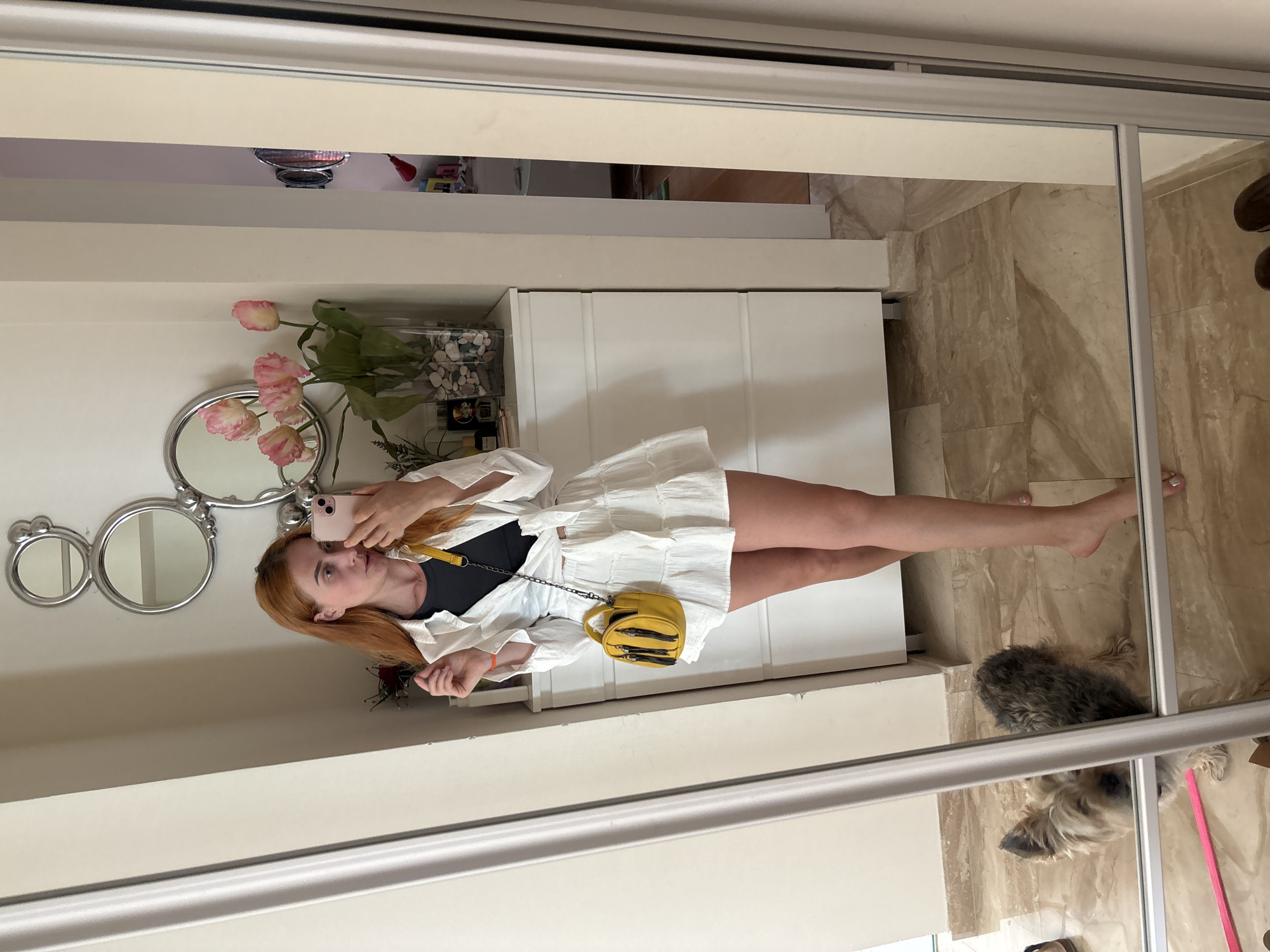 A young woman with red hair takes a mirror selfie in what appears to be a home entryway or dressing area. She is wearing a white open-collared shirt tied at the waist over a black top, and a tiered white skirt. A small, yellow, textured crossbody bag with black fringe hangs from her shoulder. She holds a pink smartphone in her right hand, angled to capture her reflection.
The background includes a tall white dresser with decorative mirrors and a vase of pink tulips above it. To the left, a closet door with a vertical handle is visible. In the lower left of the frame, a small, light brown dog with dark ears looks up towards the camera, its reflection also visible in the mirror. The floor is tiled with a marble-like pattern. In the distance, through an open doorway, a cluttered room with shelves and various items can be seen. The scene is brightly lit, suggesting it is daytime. The presence of a small dog and the domestic setting suggest this image was taken within a private residence. There is no specific indicator of the location being in the city of Budva, Montenegro, beyond the metadata that might accompany the image.