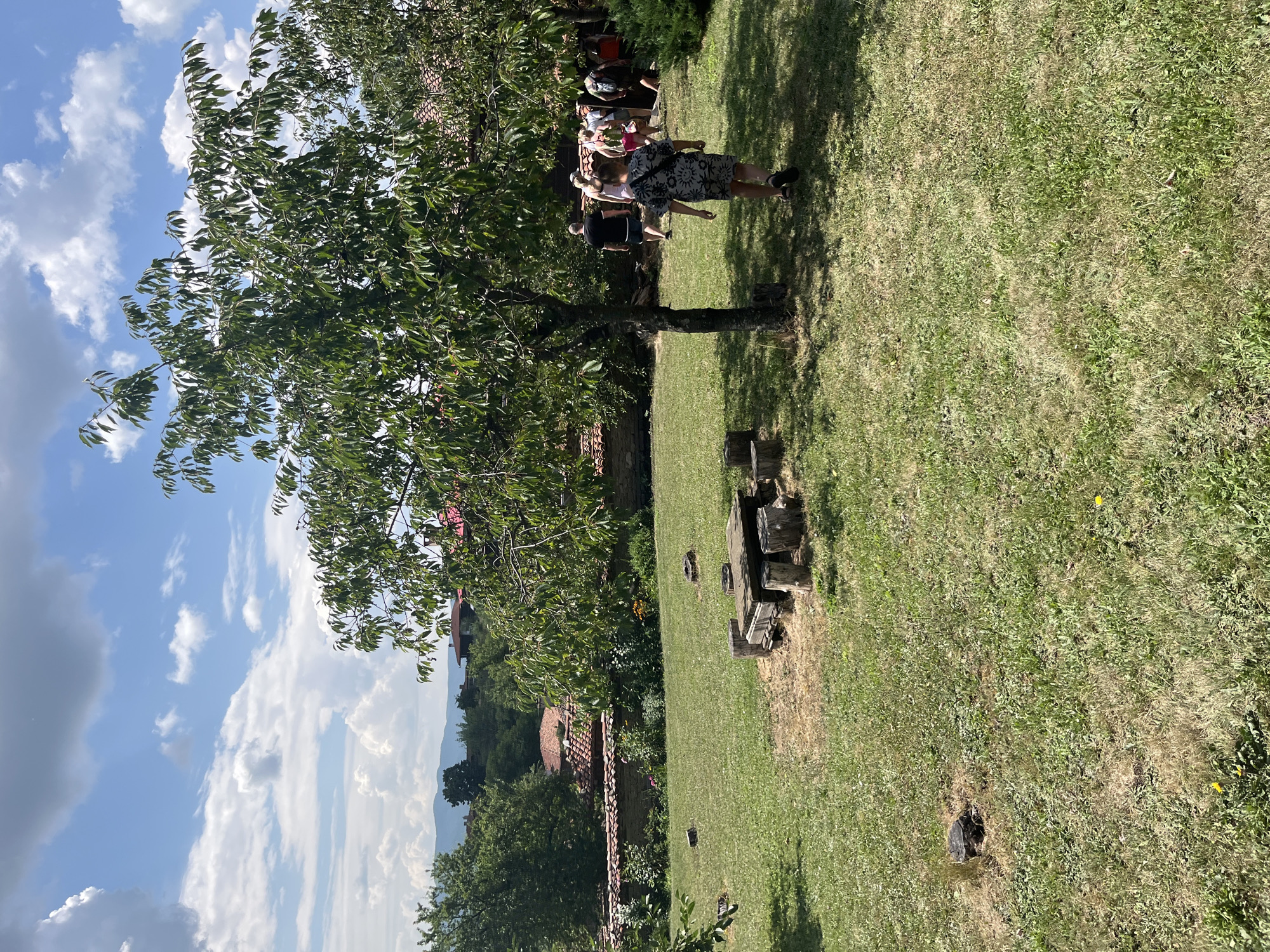 The image depicts an outdoor scene in the ethnographic complex of Zheravna, Bulgaria. The foreground features a grassy lawn with a stone table and several log seats arranged around it. A medium-sized tree with green leaves shades part of the lawn. In the mid-ground, a group of people, including adults and children, are walking away from the camera and towards buildings with traditional red-tiled roofs. The background shows more buildings, lush green trees, and a distant hilly landscape under a bright blue sky with scattered white clouds. The overall impression is a sunny day in a historic, rural setting.