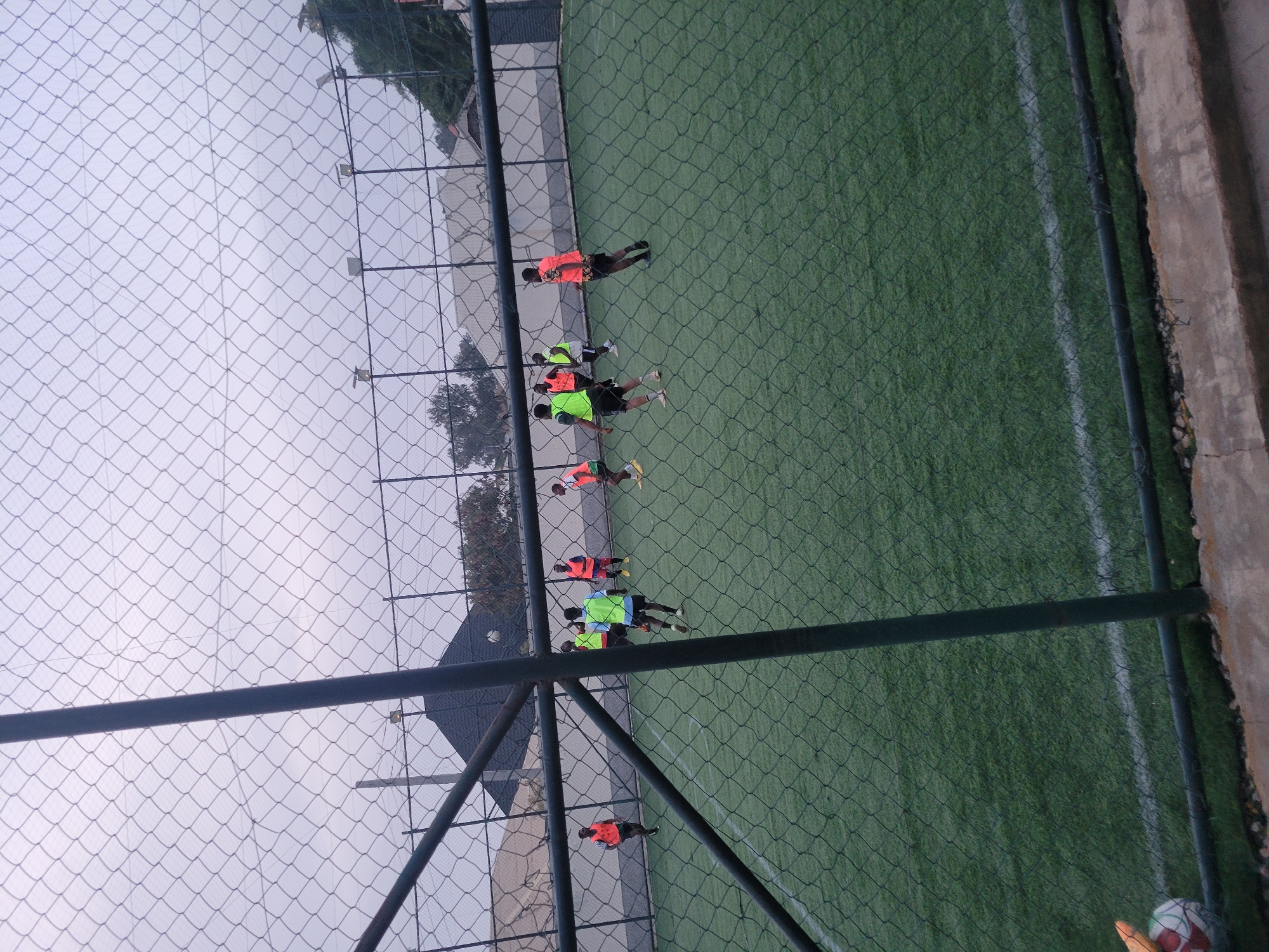 The image shows a group of people playing on an artificial turf soccer field enclosed by netting. The people are wearing bright orange and green vests. The environment includes a cloudy sky and buildings in the background. A soccer ball is visible in the foreground. The location is Unknown, Unknown.