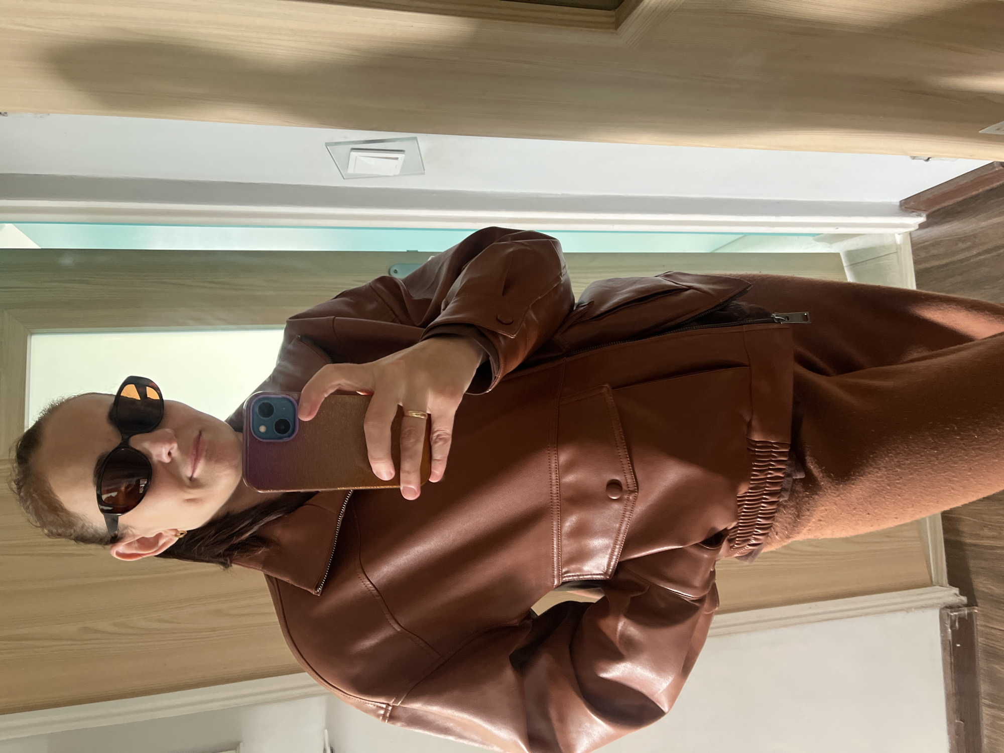A person, wearing sunglasses and a brown leather jacket, is taking a selfie with a smartphone. The background includes a doorway with white trim, light-colored wooden paneling, and a ceiling with a recessed light fixture. The location appears to be indoors, possibly a home or apartment. The city is Unknown, Unknown.