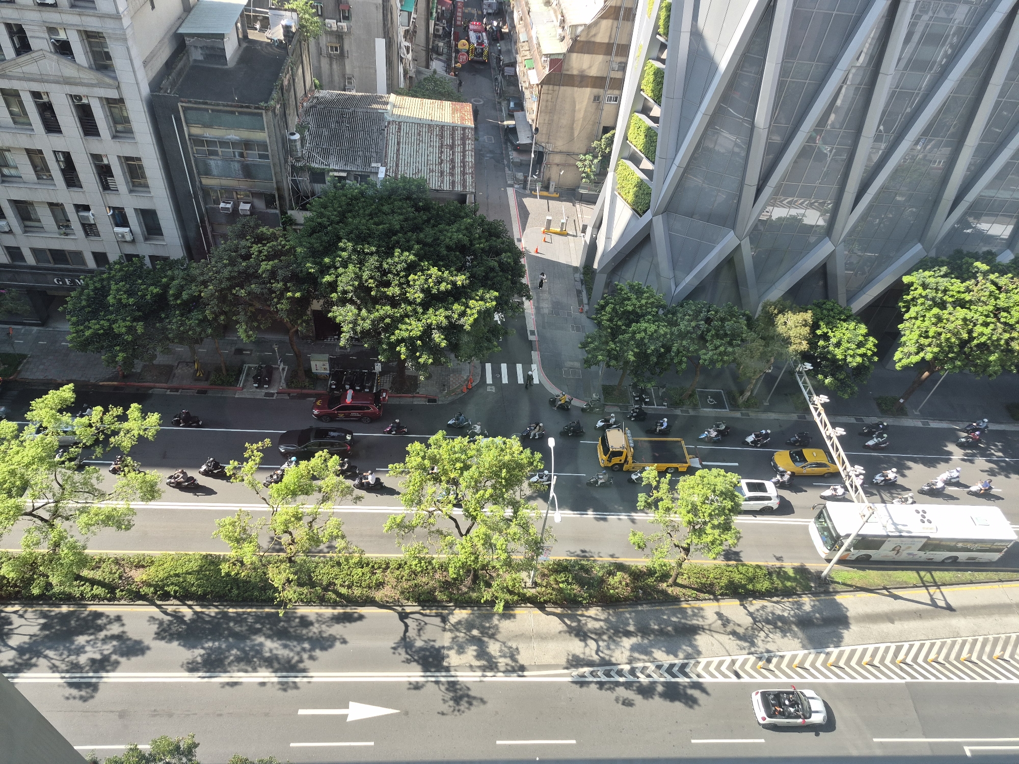 This is an aerial, high-angle shot of a street scene in Taipei, Taiwan, likely on a sunny day given the strong shadows. The street is bordered on the left by multi-story buildings, including a light-colored, modern building with many windows and a sign that reads "GEM." Adjacent to it is an older, darker building with visible air conditioning units. On the right side of the street, a strikingly modern, angular building with a glass facade and integrated green vertical gardens dominates the scene.
The street itself is a busy thoroughfare with multiple lanes of traffic. There are numerous motorcycles and scooters moving in both directions, as well as a few cars and a white bus. A yellow flatbed truck and a yellow taxi are also visible. Pedestrians are seen crossing the street at designated crosswalks. Trees with lush green foliage line the sides of the road, casting dappled shadows on the pavement. The overall impression is one of a dynamic urban environment with a mix of older and newer architectural styles and active city life.