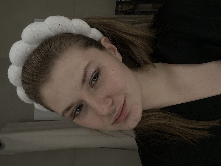 A young woman with fair skin and light brown hair pulled back in a ponytail poses at an angle. She wears a white, puffy headband. Her makeup includes a light shimmer on her eyelids and darkened lashes. She has a slight, subtle smile. She wears a black top and is partially in shadow. The background shows a blurred glimpse of a white surface and possibly a shelf with items.