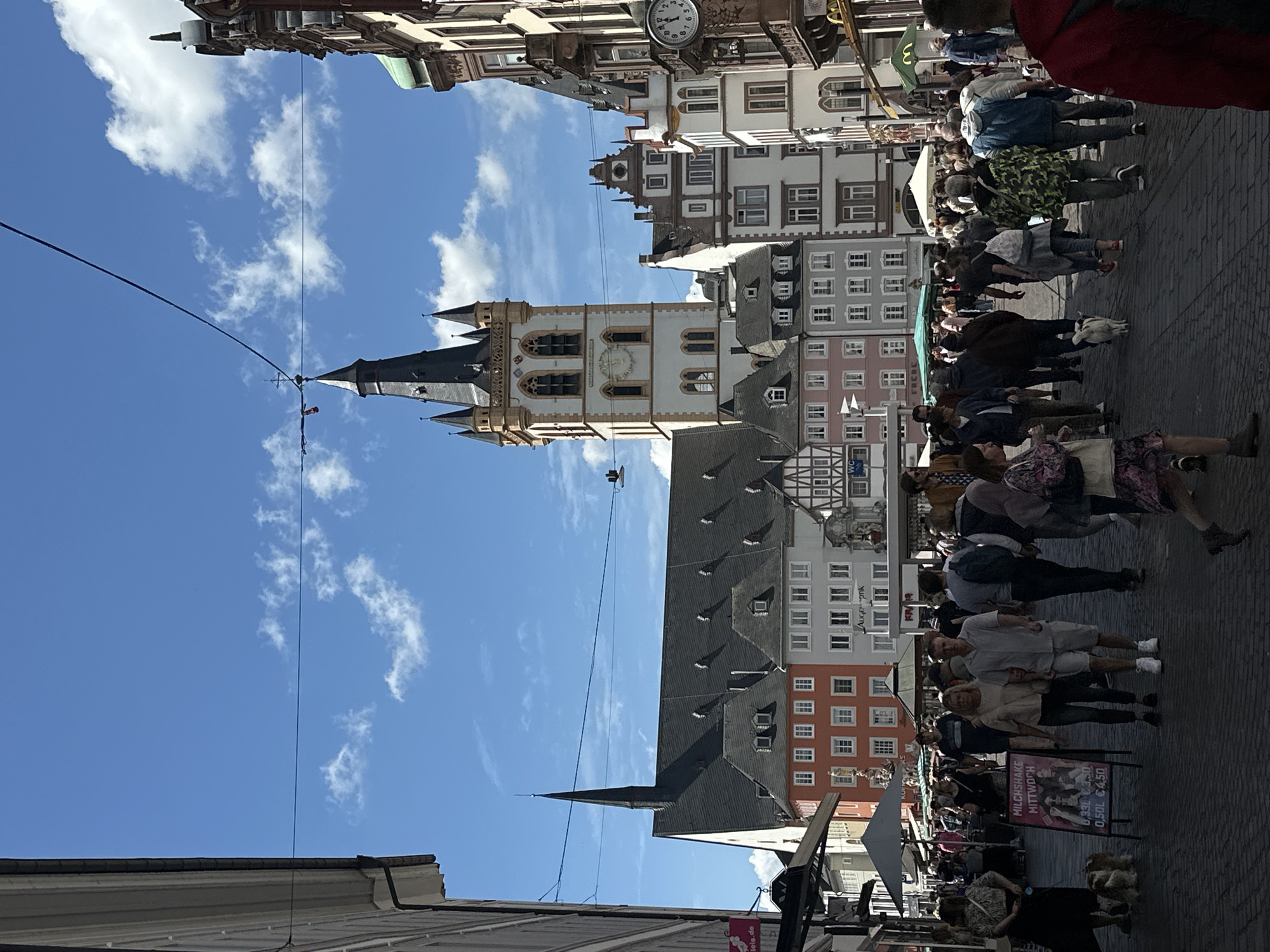 This detailed image captures a bustling pedestrian street in Limburg an der Lahn, Germany, under a clear blue sky with scattered white clouds.
In the background, dominating the right-of-center skyline, is the Werner-Senger-Haus tower, part of the Limburg Town Hall. It is a tall, ornate structure with a white or cream-colored facade, a prominent dark pointed roof, and several smaller spires or turrets. A clock face is visible on one side of the tower. To the left of this tower, further in the background, the Limburg Cathedral (Dom St. Georg) is partially visible, characterized by its extensive, steeply pitched dark roof and a slender spire on its left.
The street is lined with multi-story historic buildings exhibiting a variety of architectural styles. Many feature colorful painted facades in shades of red, pink, grey, and white, with numerous windows and gabled roofs, some including dormer windows. Half-timbered elements are visible on some structures. On the far right, a building has a section with a green domed or sloped roof. A circular clock is mounted on a building facade on the right side of the street, indicating the time is approximately 6:25. Overhead, multiple thin black wires crisscross the sky, strung between buildings, with some supporting street light fixtures. A white "WC" sign is visible on a building to the far left.
The foreground and midground are filled with numerous people walking in various directions along a cobblestone street, suggesting a busy market or shopping area. Most individuals are casually dressed. In the immediate foreground, slightly left of center, a large A-frame sign stands on the pavement, advertising "MILCHSHAKE MITTWOCH" (Milkshake Wednesday) with prices listed as "0,33L €3.50" and "0,50L €4.50," along with images of milkshakes. To the left of this sign, a woman stands near two small, fluffy, light-colored dogs. In the mid-ground, a couple walks arm-in-arm towards the viewer; the woman has blonde hair and wears a light top and dark pants, while the man wears a light short-sleeved shirt and light shorts. Several other individuals are seen walking with dogs on leashes. On the far right edge of the image, the back of a person wearing a red hoodie or jacket is visible. Further in the background, market stalls with awnings or umbrellas are set up along the street.