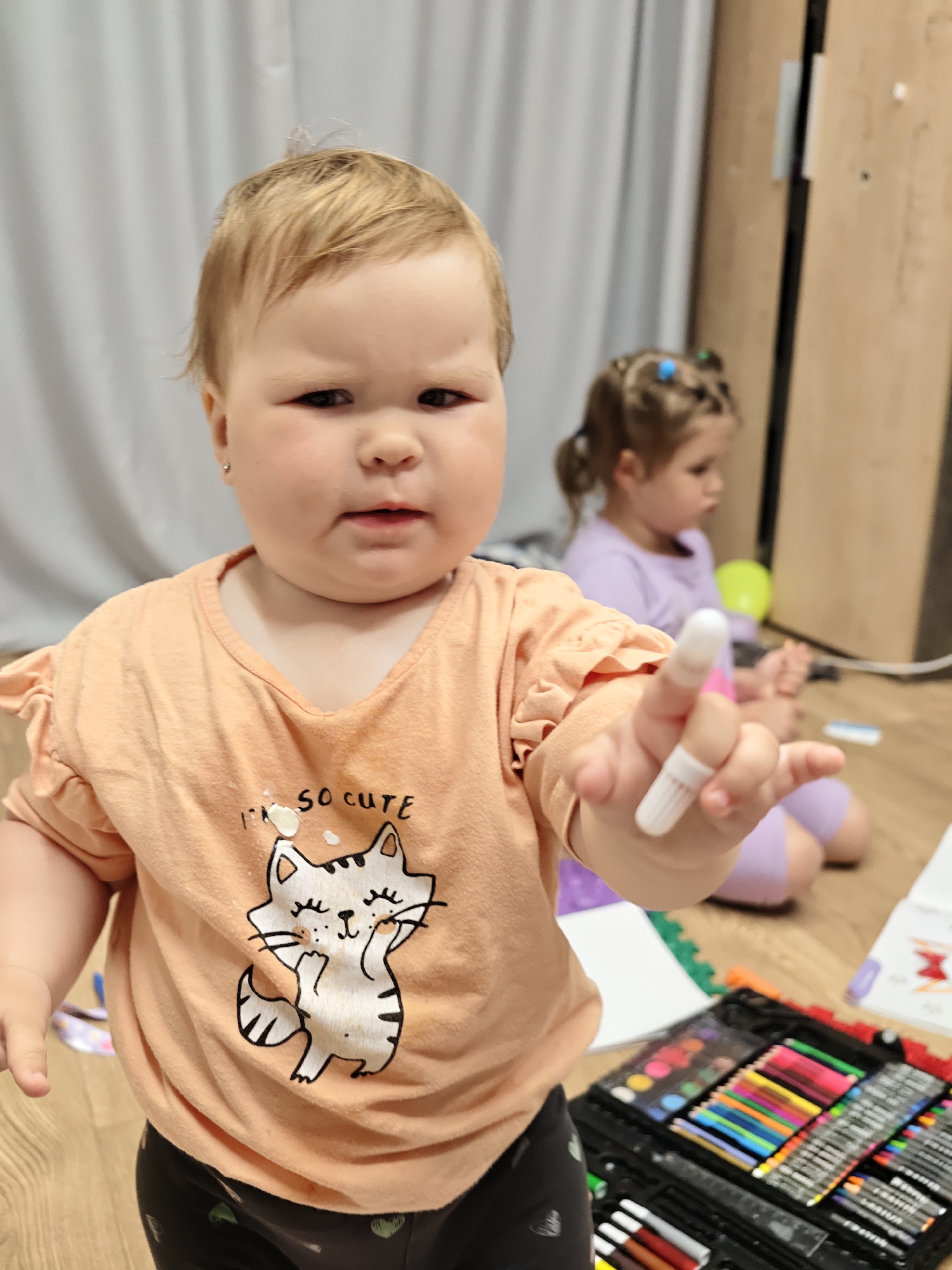 A toddler with blonde hair and a light orange t-shirt with a cat graphic is holding a pink marker. The child has a displeased expression. In the background, another toddler with brown hair in pigtails sits on the floor. The environment appears to be an indoor setting with light-colored curtains and wooden flooring. A case of art supplies, including markers and colored pencils, is visible in the foreground. The scene is likely a home in the city of Chaikovsky, Russia.