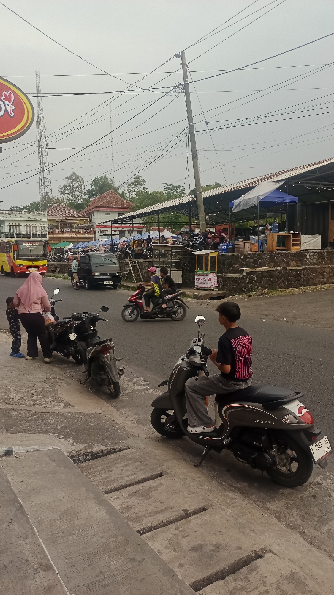 The image depicts a street scene in Pakemitan, Indonesia, characterized by a somewhat overcast sky. The foreground shows a concrete roadside with several parked motorcycles. A person wearing a pink hijab and a child in patterned clothing stand near these motorcycles. Further back, a black van is parked on the asphalt road.
Across the street, a red and yellow bus is visible. In the background, there are several open-air structures with pitched roofs, some with blue awnings, suggesting commercial establishments such as food stalls or shops. People are seen seated at tables and some are on motorcycles. A power pole with numerous wires is a prominent feature. The overall environment appears to be a busy, informal commercial area.