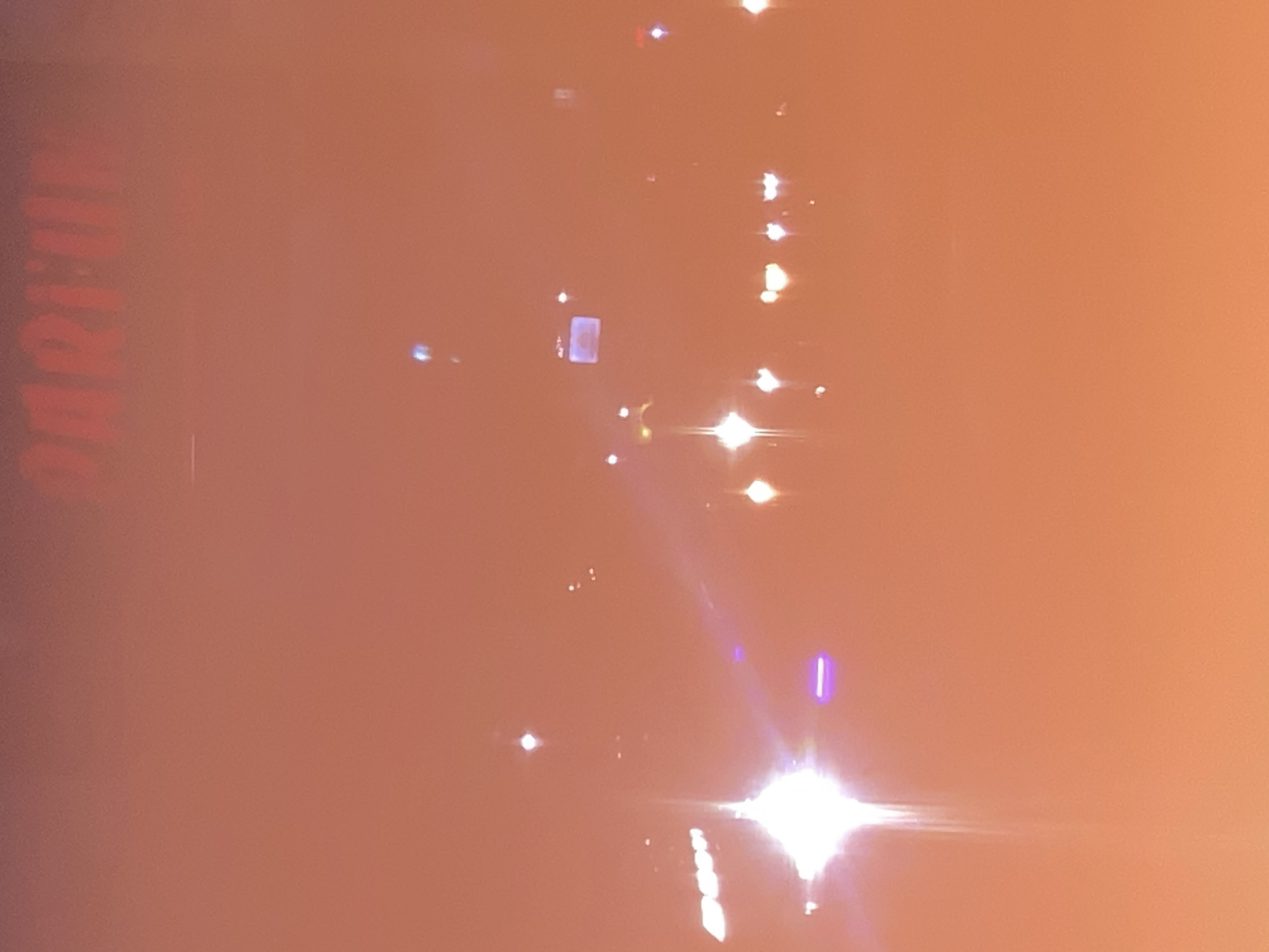 The image shows a blurry, orange-hued scene with bright lights and indistinct shapes, suggesting it was taken at night or in low light conditions. At the top of the image, red lettering appears to spell out "PARFUM," possibly indicating a sign for a business. Several points of light are scattered throughout the frame, some appearing as starbursts, likely from streetlights or vehicle headlights. There are no visible people or identifiable structures that would clearly place the location in Kasihan, Indonesia. The overall impression is of an out-of-focus urban environment at night.