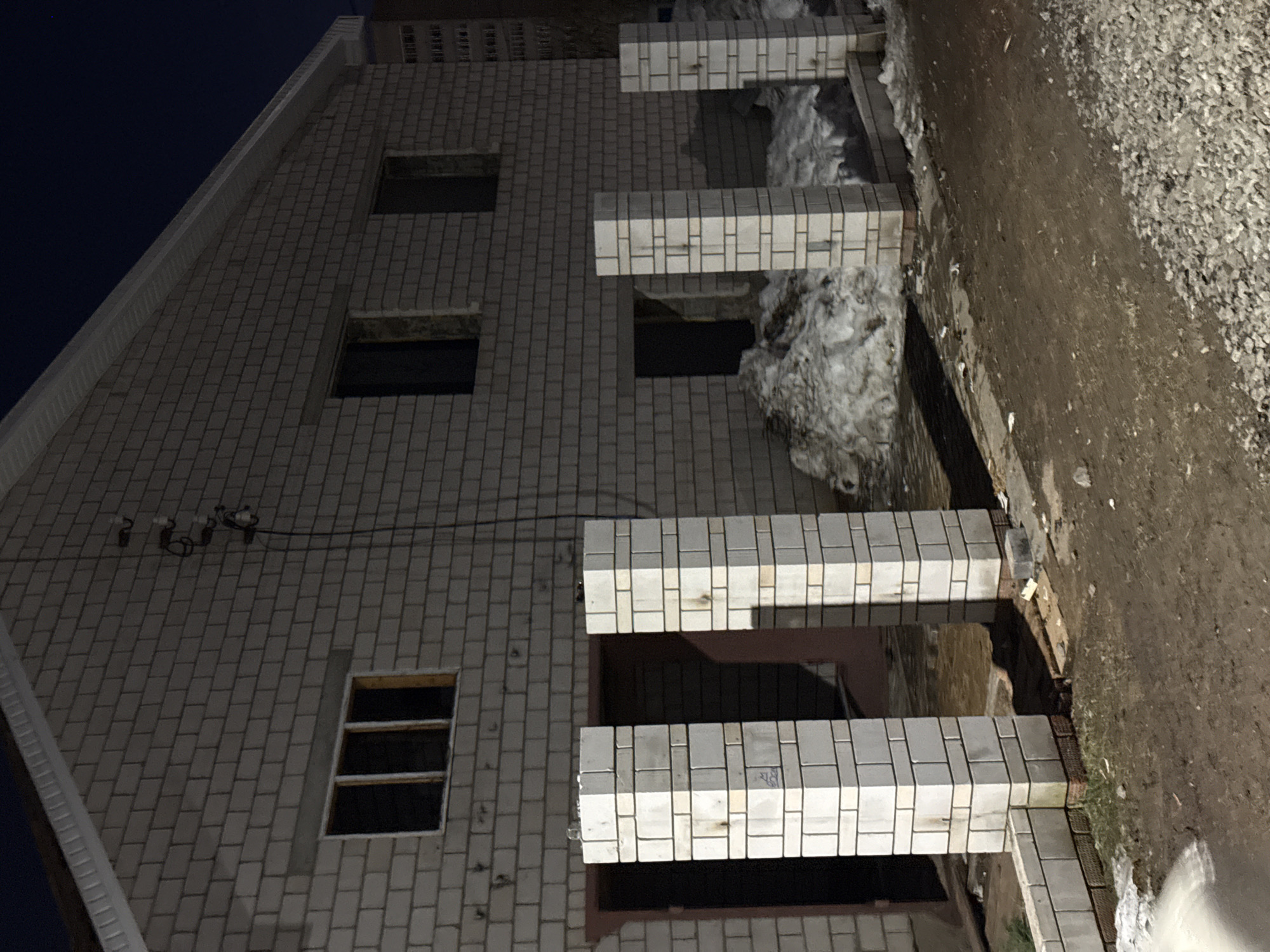 The image shows a multi-story brick building under construction at night or in low light. The building appears to be located at the periphery of the city of Izhevsk, Russia. The building's exterior is primarily white brick, and it features multiple rectangular windows and what appear to be unfinished balcony structures with brick columns. Some areas show patches of snow or ice accumulation, and the ground adjacent to the building consists of dirt and gravel. Electrical cables run along the exterior wall near the roofline. The scene is undeveloped, likely on the edge of the developed zone of the city. The building appears incomplete, suggesting it is still under construction. No people, objects, or actions can be observed.