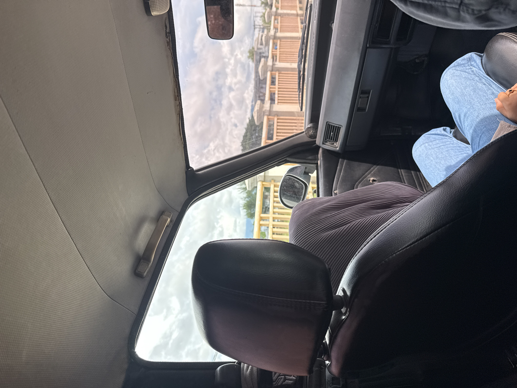 The image shows the interior of a car, looking out the windshield. The dashboard and part of the passenger seat are visible. A person wearing light blue jeans is seated in the passenger seat. Outside the car, a cloudy sky is visible, along with a light brown fence and some buildings. The scene appears to be in Glameunasah Baro, Indonesia.