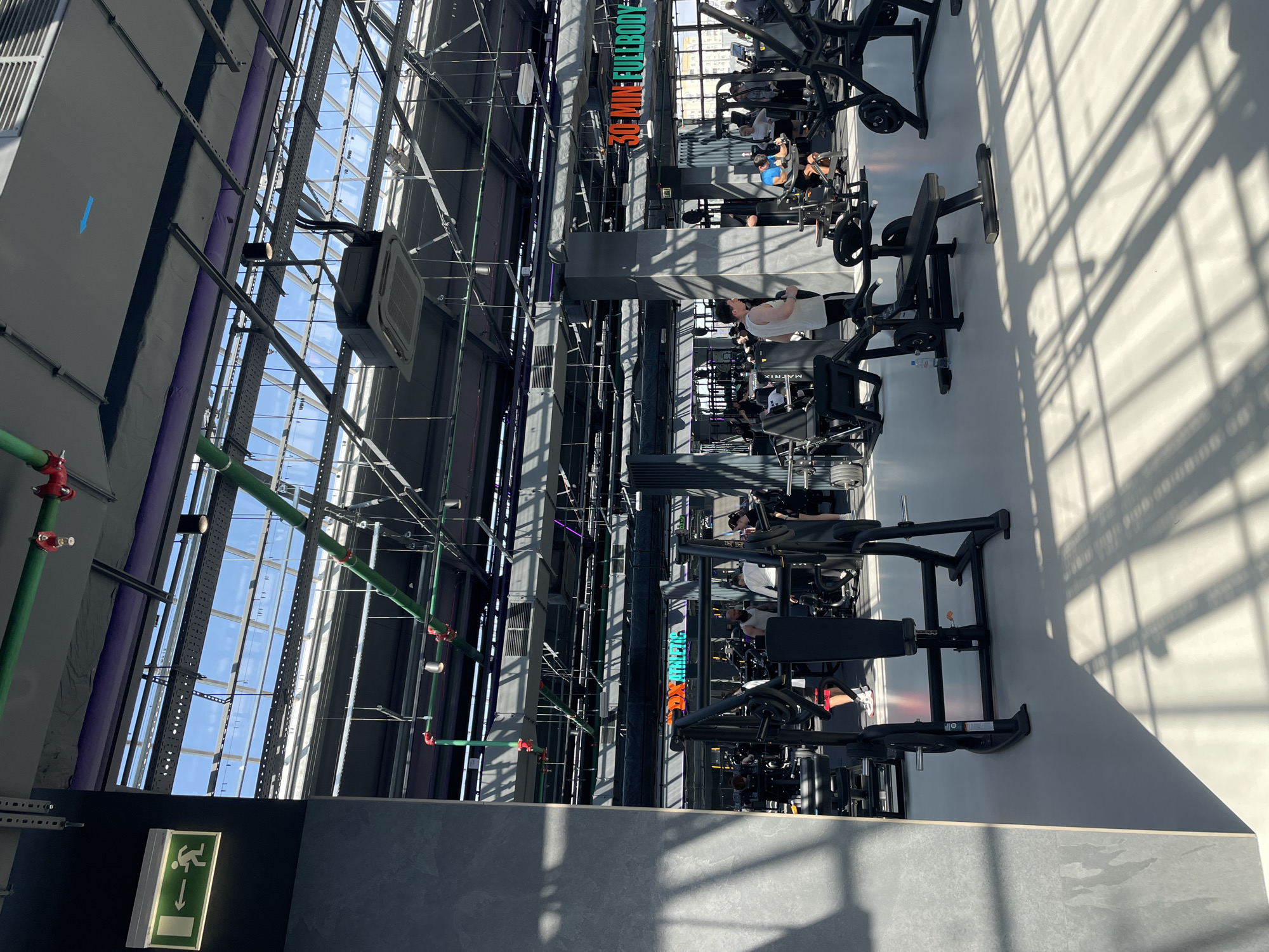 The image shows the interior of a gym in Kazan, Russia. The gym has a high ceiling with a glass roof, supported by metal beams. There are several exercise machines arranged in rows. People are using the equipment. There are signs with "30-Minute Fullbody" in orange and green. There are green and red pipes running along the ceiling and walls. An emergency exit sign is visible in the lower-left corner. Sunlight streams through the glass roof, casting shadows on the floor.