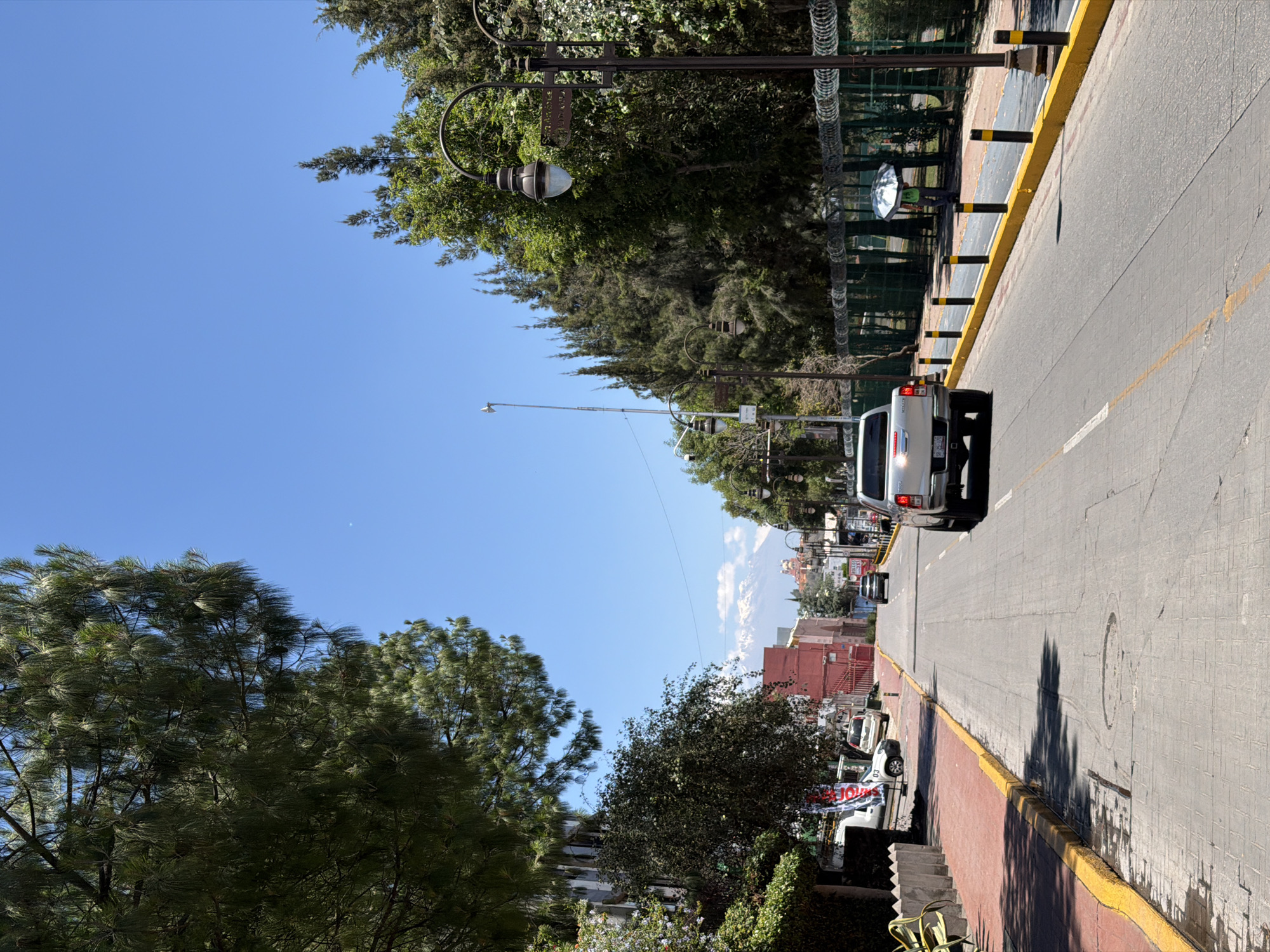 The image depicts a street view in San Andrés Cholula, Mexico, under a clear blue sky. The road is paved with rectangular stones and has a yellow line marking the shoulder. A silver pickup truck is driving away from the viewer on the right side of the street. On the right side of the street is a fence with barbed wire on top of it. There are black and yellow vertical bollards along the sidewalk. On the left side of the street, a flight of stairs is visible, leading up to an area with shrubs and greenery. Several buildings are seen in the distance, with one painted red. The image shows trees on both sides of the road, and streetlights are mounted on lampposts along the right side. Banners are hanging on the fence.