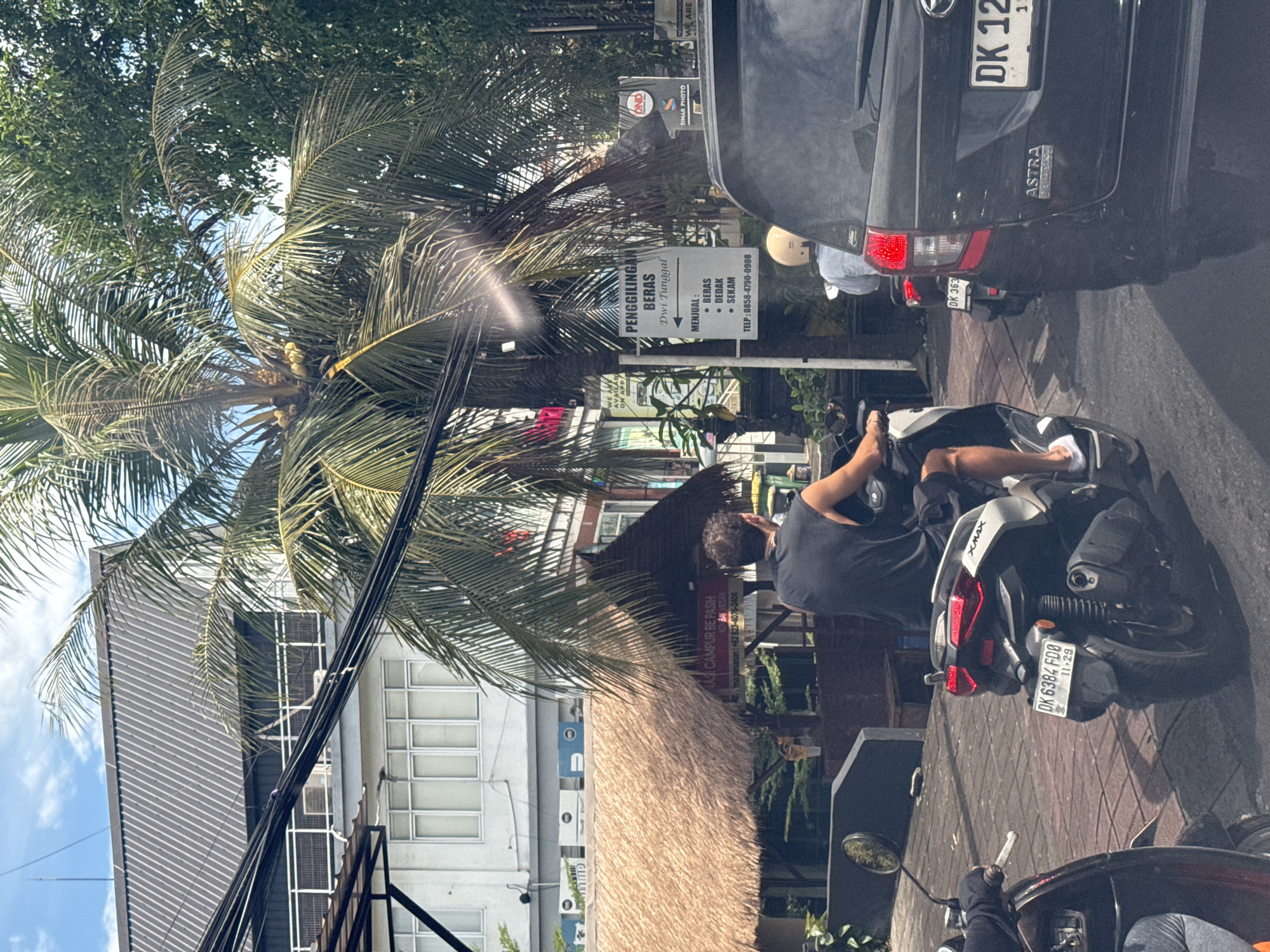 The image captures a street scene in what appears to be North Kuta, Indonesia. A man is seated on a black and white XMAX scooter, license plate DK 6384 FDO. Another scooter is parked behind his, and a black Astra vehicle, license plate DK 12. A man with a helmet is riding near the black car. Signs for “PENGGILINGAN BERAS Dwi Tunggal” (rice milling) and "CAMPUR BE PASIH" (a restaurant) are visible behind the scooter and riders. The rice milling sign indicates that it sells rice, bran, and husk. A tall palm tree and building are located behind the sign. Overhead electrical wires stretch across the top of the image. The environment appears to be urban, with evidence of business activities and traffic.