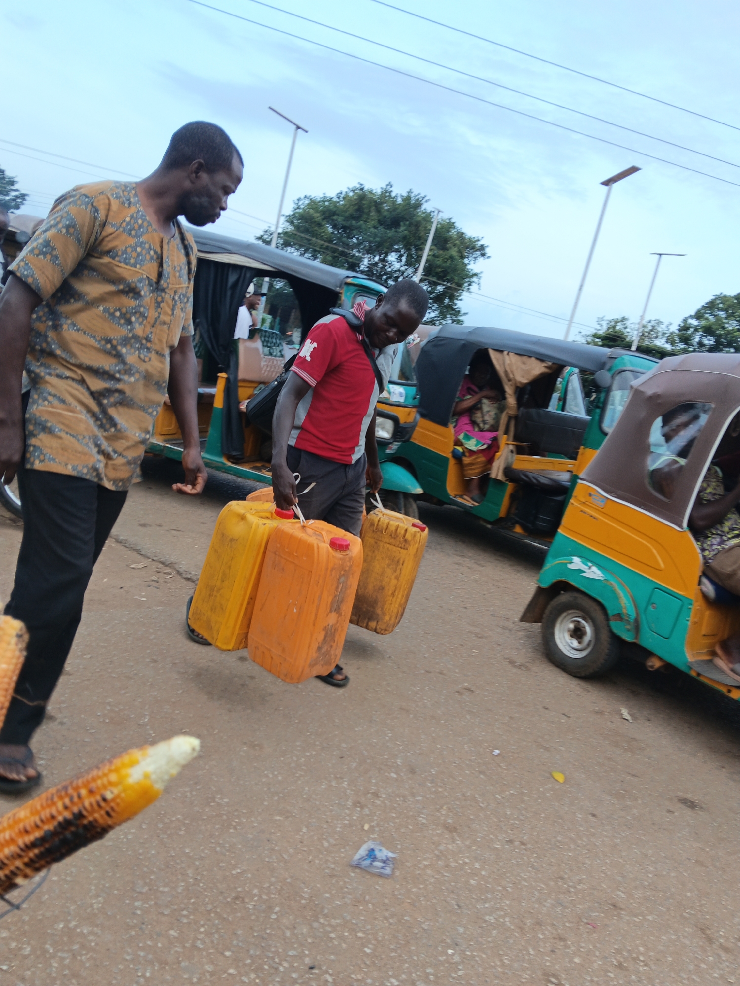 In a street scene under a cloudy sky, two men are carrying large yellow and orange plastic jerrycans. The man on the left wears a patterned shirt and black pants, while the man on the right wears a red shirt and dark shorts. They are walking next to two auto-rickshaws. The auto-rickshaws are green and yellow with brown canopies, and people are visible inside them. In the foreground, a roasted ear of corn is positioned close to the camera. The ground is paved with asphalt and scattered with small debris.