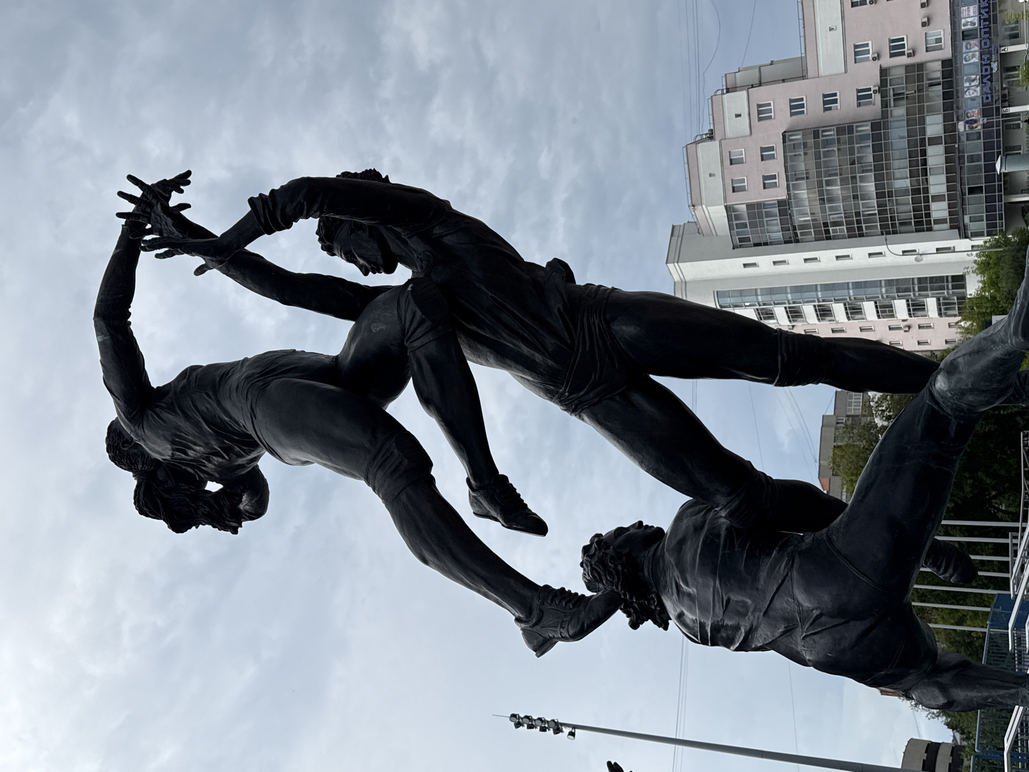 A dark bronze sculpture depicts three male figures in athletic poses, stacked vertically. The bottom figure is partially visible, with its head and torso angled away from the camera. The middle figure supports the top figure, its body contorted as it reaches upwards. The top figure is in mid-air, legs bent and arms extended, seemingly in a moment of acrobatic action. All figures are clad in what appears to be athletic attire, including shorts and shirts.
In the background, a modern, multi-story building with a pink and white facade, featuring numerous windows and balconies, rises against a cloudy sky. To the left of the sculpture, a tall metal pole with lights is visible, along with trees and other urban structures. The sculpture is situated in Yekaterinburg, Russia, likely in a public space.