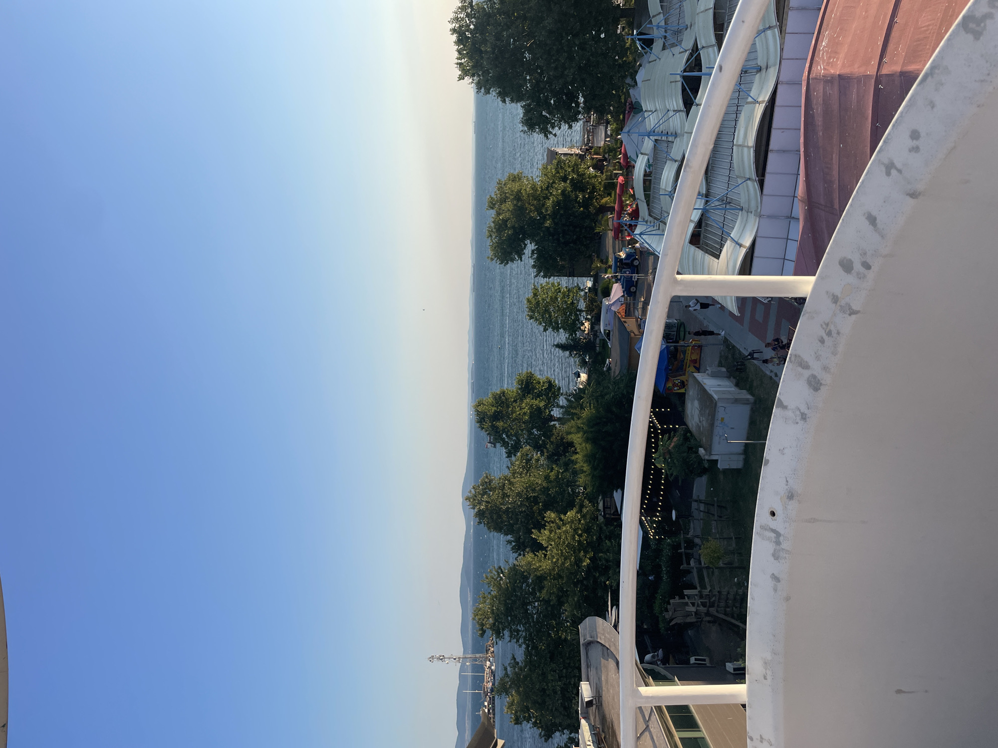 The image captures a scenic view of the Black Sea from a high vantage point, likely a hotel or a public building, in Pomorie, Bulgaria. The foreground is dominated by a curved, white railing and a portion of a white structure, suggesting a balcony or observation deck. Below, a cluster of buildings with red and white roofs, possibly market stalls or small shops, are visible. Green trees line the coastline, separating the urban area from the sea. The sea itself is a deep blue, with several boats and ships dotting the horizon. In the distance, a chain of low hills forms the background under a clear, pale blue sky. The overall impression is one of a tranquil coastal town on a sunny day.