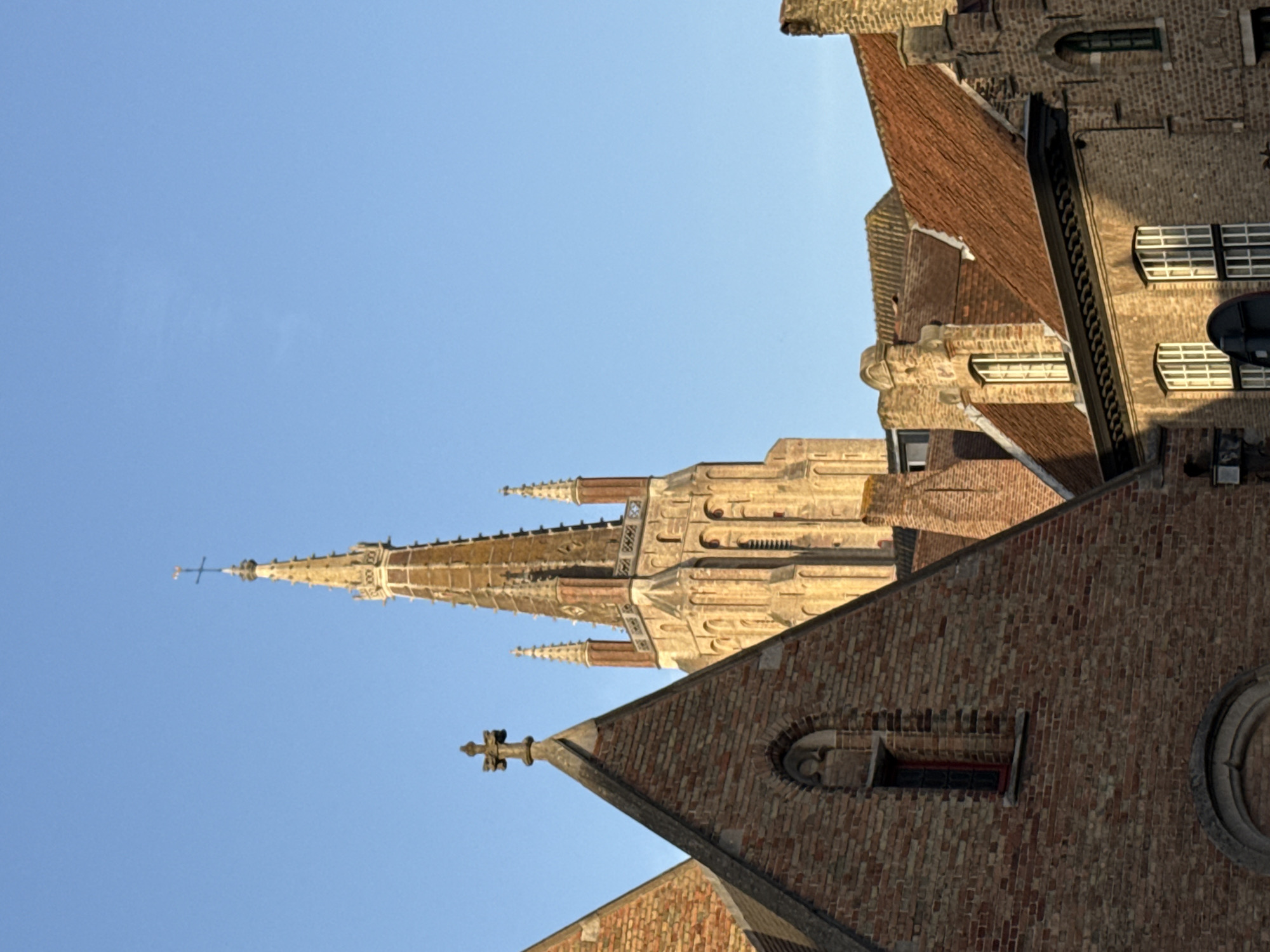 This is a photograph of a church spire and surrounding brick buildings under a clear blue sky. The perspective is from a low angle looking upwards. The church spire is tall and pointed, constructed of stone with ornate architectural details, including what appears to be small spires and decorative elements along its length. It is topped with a cross.
The surrounding buildings are made of red brick and have red tile roofs. One building in the foreground has arched window openings with decorative stonework. Another building, further back and to the right, also features brickwork and windows. The architectural style suggests a European, possibly medieval or Gothic, influence, consistent with the location context of Bruges, Belgium.
The sunlight suggests it is daytime, likely morning or late afternoon, given the warm tones on the brickwork and spire. The clear blue sky indicates fair weather. There are no people visible in the image. The focus is on the architecture and the grandeur of the church spire against the sky. No visible text is present.