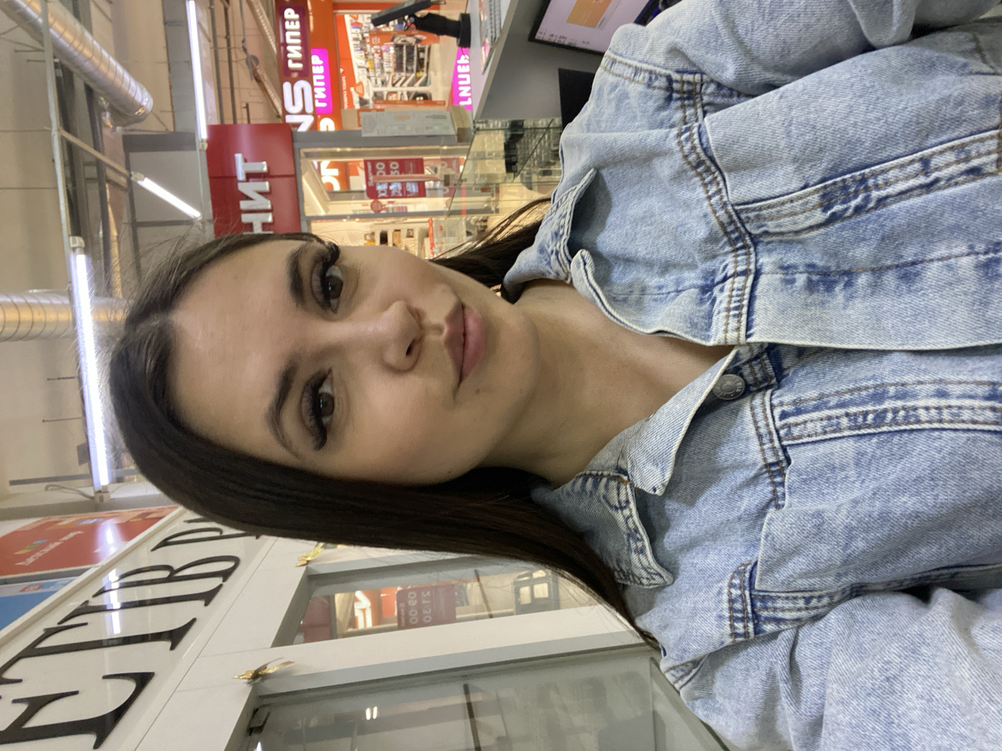 The image shows a woman wearing a light blue denim jacket, indoors, likely in a shopping mall in Chaikovsky, Russia. Store signs with Russian writing are visible in the background, including the word "ГИПЕР", which means "Hyper". There's a display screen visible, and some store fronts behind a glass barrier.