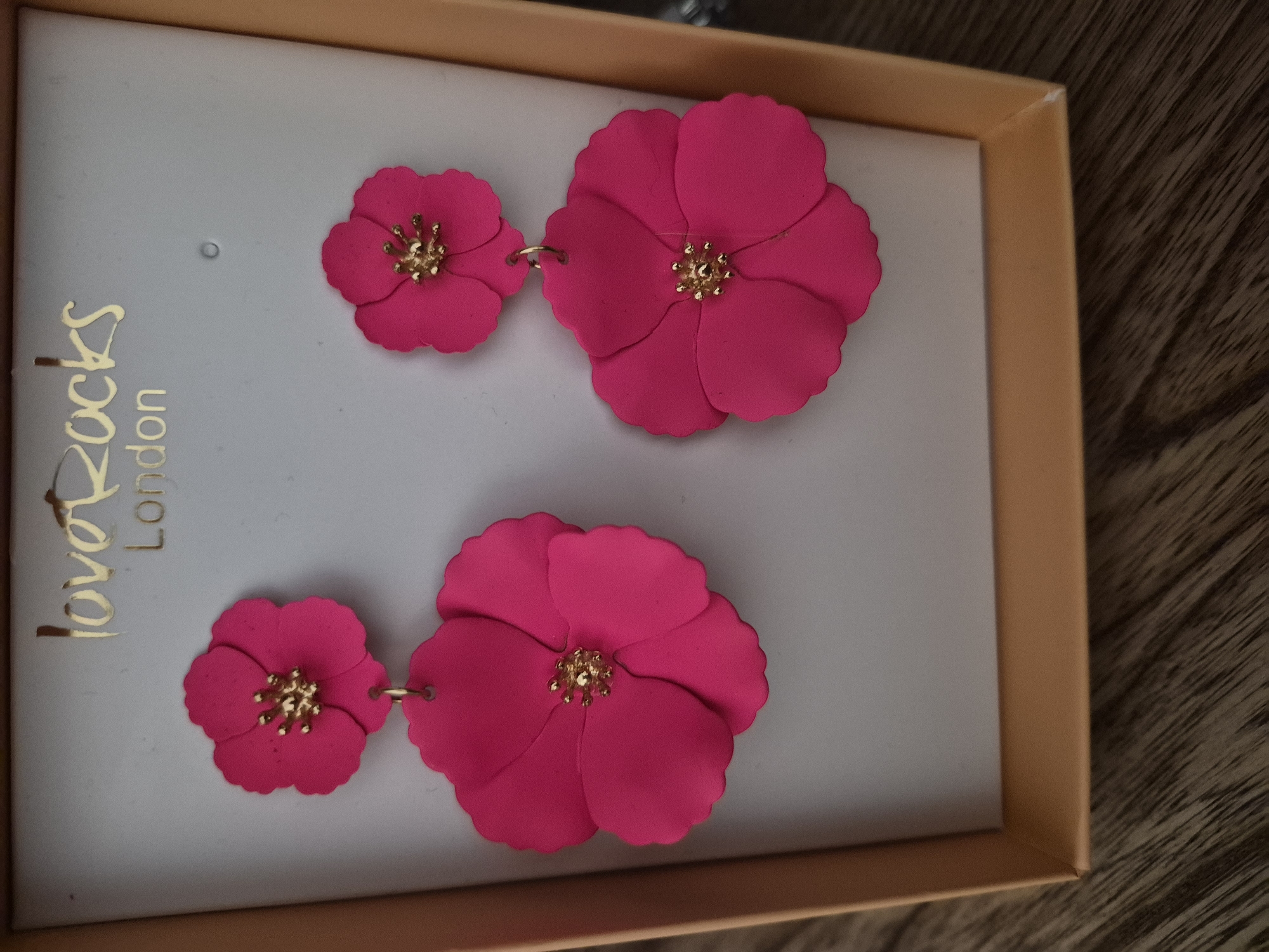 An overhead medium shot shows a pair of fuchsia-pink floral drop earrings with gold-tone centers, displayed in their original peach-colored box. The earrings are attached to a light gray cardboard insert within the box, which also features a gold-tone logo that reads "love Rocks London." The box itself is open, revealing the bottom edge of the display and a portion of a dark brown wooden surface below.