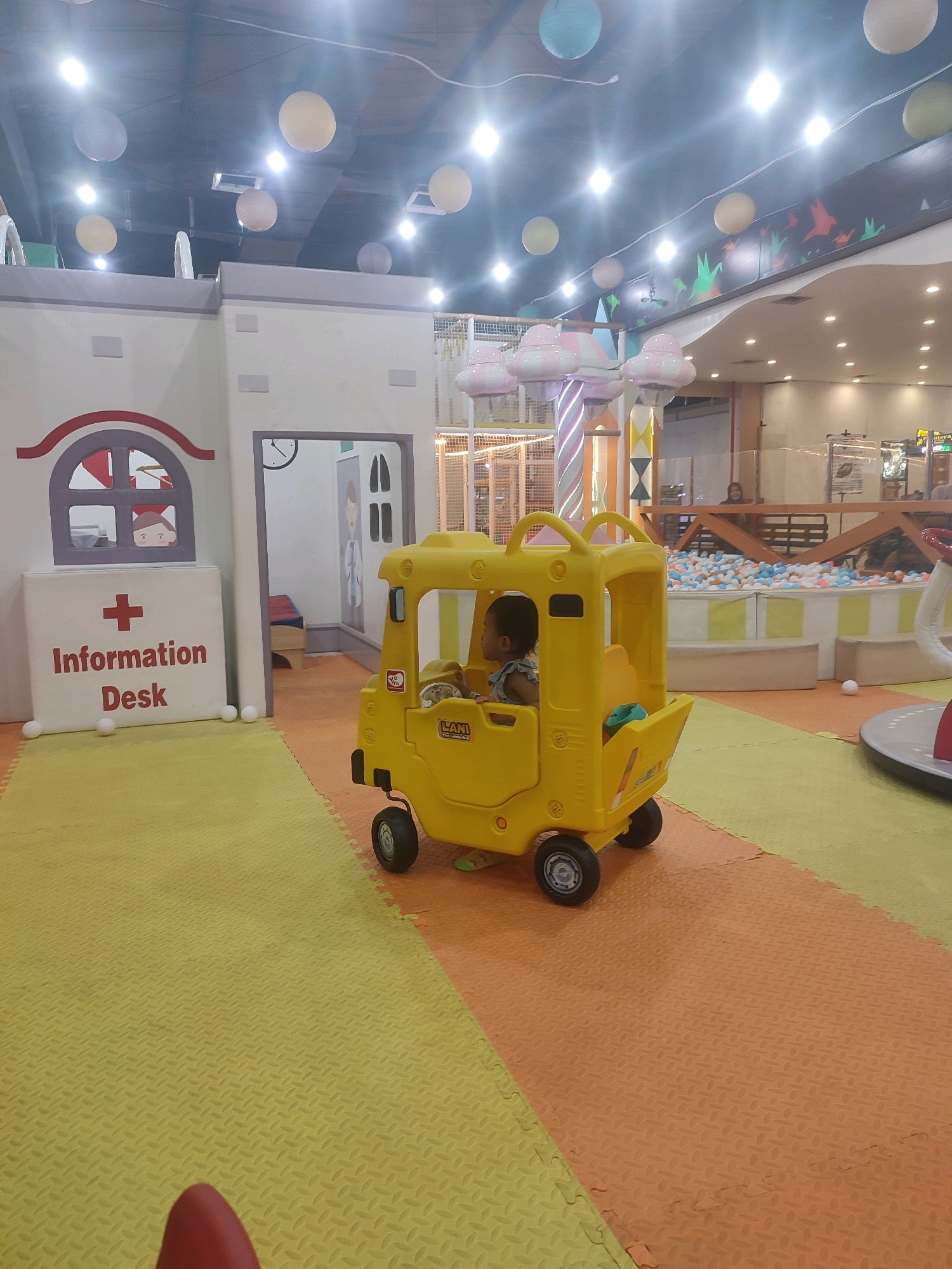 The image depicts an indoor children's play area, likely in Kediri City, Indonesia. In the foreground, a young child sits in a yellow toy bus. The bus has a clear plastic windshield and a steering wheel. The child is looking to the left. The floor is covered with interlocking foam mats in yellow and orange.
Behind the toy bus, there is a mock "Information Desk" with a red cross symbol and the words "Information Desk" in red letters. Above the desk is a window depicting a cartoon character. To the right of the information desk is an open doorway leading into another room, which appears to be a playhouse with some furniture.
In the background, there is a ball pit filled with white, blue, and orange balls, and a section of play equipment with colorful vertical stripes and what appear to be cloud-shaped decorations hanging from the ceiling. The ceiling is dark with numerous bright lights, and several round, translucent paper lanterns are suspended from it. The overall impression is that of a lively and safe indoor play environment for young children.