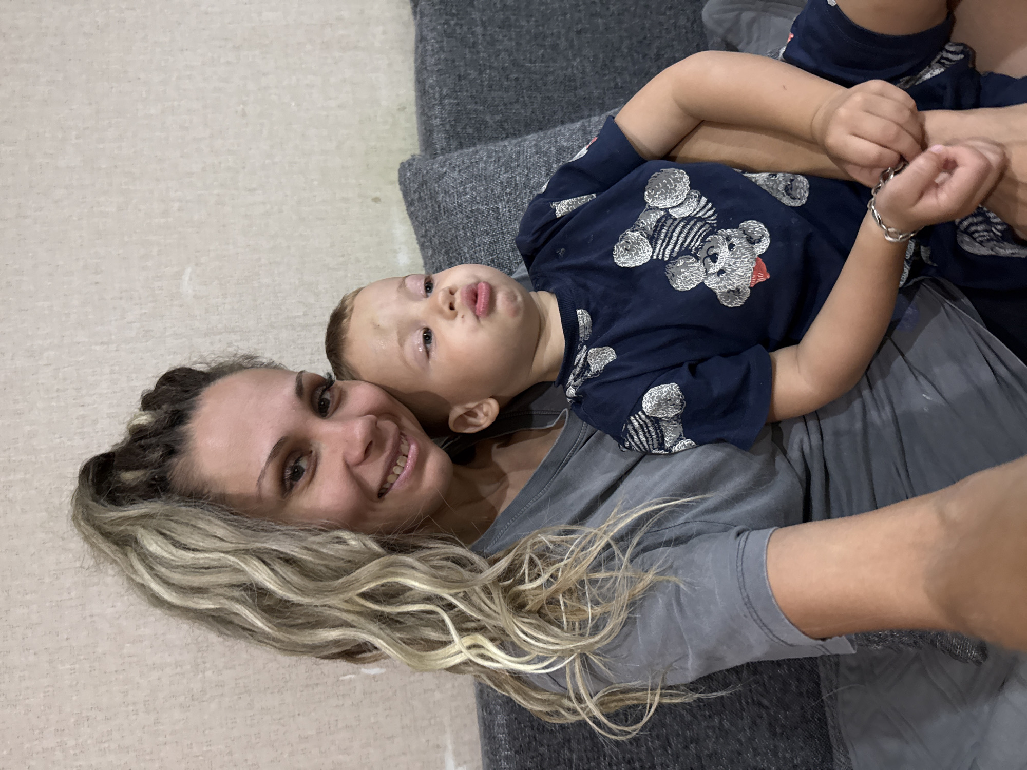 A woman with blonde and brown braided hair, wearing a grey t-shirt, is sitting on a grey sofa and holding a young child on her lap. The child, wearing a dark blue t-shirt with a teddy bear pattern, is looking to the right and has their lips puckered. The background appears to be a wall with textured wallpaper. This scene is set in a residential location in Rusanovo, Russia.