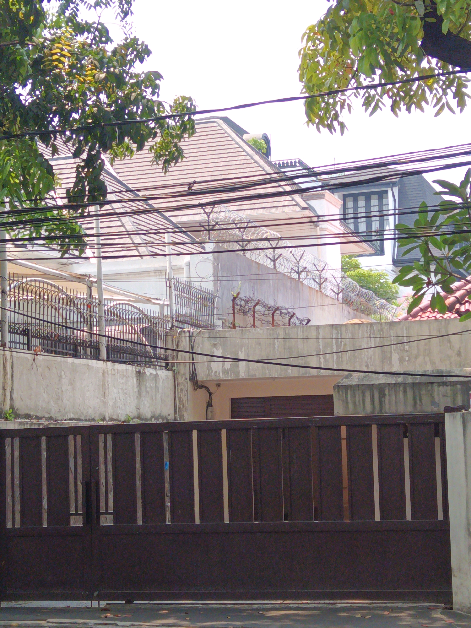 A medium shot captures a street-level view of a secured property in Central Jakarta, Indonesia, dominated by a large, dark brown metal gate with vertical slats. Beyond the gate, a concrete wall topped with a decorative black metal fence encloses the property. Further in, a building with a light-colored facade and a brown tiled roof is visible, partially obscured by trees. Another building with a dark gray roof and windows is also present. Barbed wire is installed along the top of the outer wall, indicating enhanced security. Electrical wires crisscross the scene in the foreground and midground. The environment appears to be a residential or commercial area with substantial security measures.