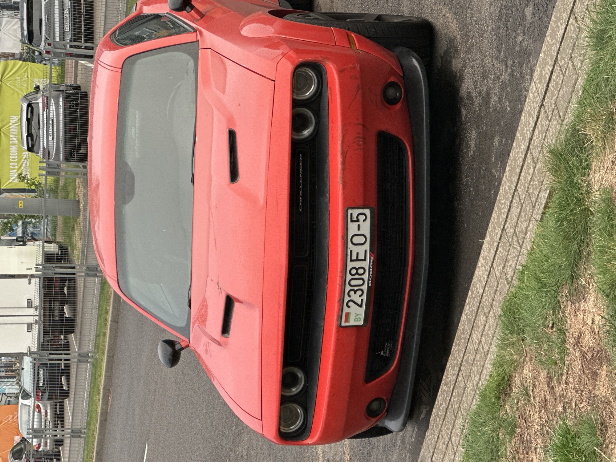A red Dodge Challenger, bearing Belarusian license plate BY 2308 EO-5, is centrally positioned, parked on a wet asphalt surface. The vehicle exhibits water spots across its body and minor scuffing on the lower portion of its front bumper. A concrete curb separates the asphalt from a narrow strip of grass and dry earth in the foreground. In the midground, beyond the Dodge Challenger, a metal fence separates the current parking area from another parking lot. Behind this fence, multiple vehicles are visible, including a silver sedan, a dark SUV with Belarusian license plate 2190 FI-5 (partially visible), a large white truck, and a silver SUV with Belarusian license plate 5769 MO-7. In the far background, behind the vehicles and fence, a large yellow banner is present, displaying Cyrillic text which includes "ДОМА СО СВОИМ ПАРКИНГОМ" and "НОВАЯ БОРО". The text "НОВАЯ БОРО" (New Borovaya) specifically places the scene within the Бараўлянскі сельскі Савет (Barawlyanski Sieĺski Saviet) in Belarus. The scene is illuminated by overcast conditions. No individuals are visible.