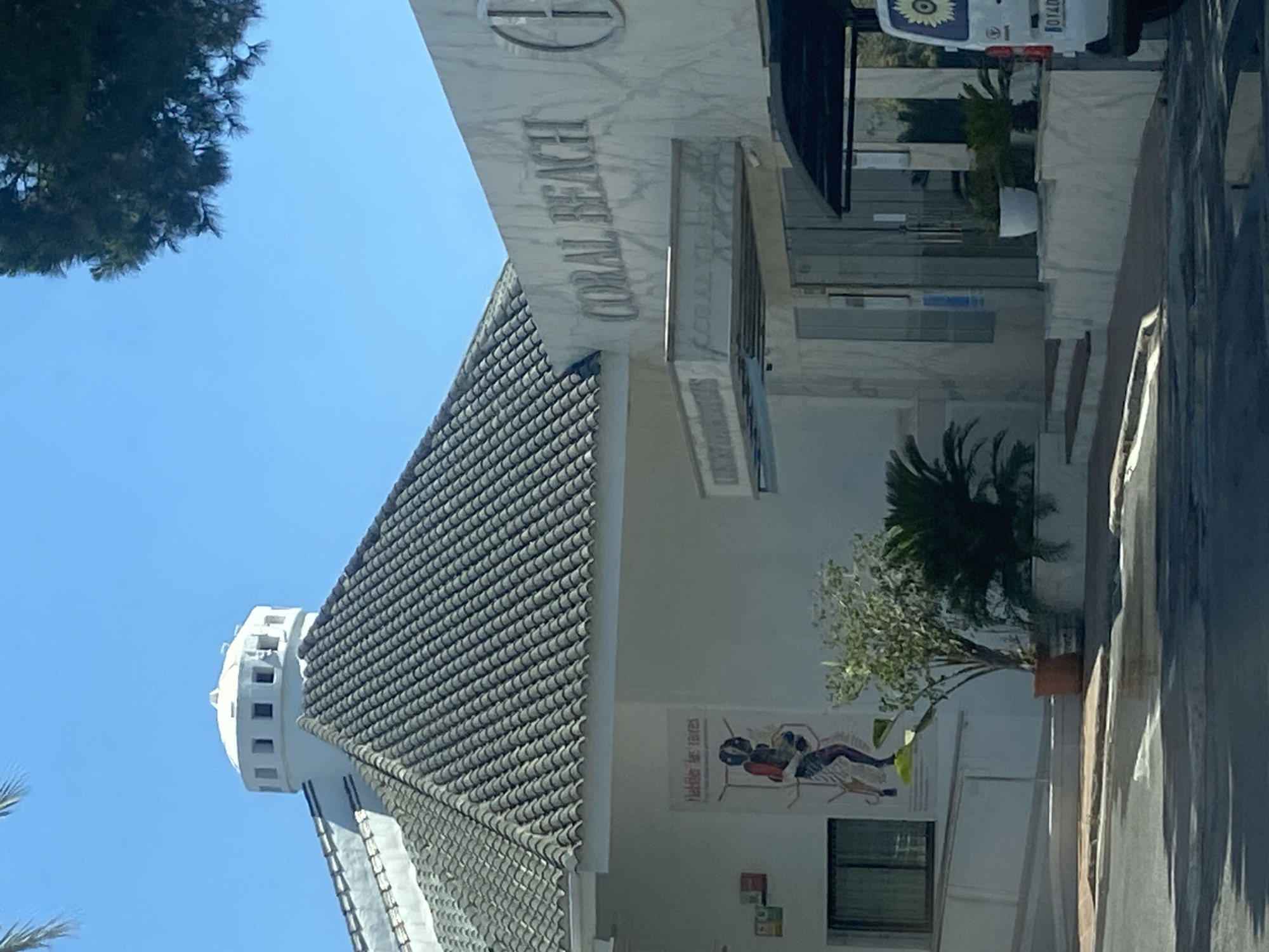 The image shows a building with "CORAL BEACH" inscribed on its facade, indicating it is likely a hotel or resort in Marbella, Spain. The building features a white marble exterior and a distinctive tiled roof. To the left, a tower-like structure with a round top and rectangular windows rises against a clear blue sky, suggesting a sunny day.
In front of the building, there is a potted tree with green foliage, and a poster or artwork is displayed on the wall. The poster depicts stylized figures, possibly in an artistic or informational context. The overall scene appears to be outdoors, with a paved walkway or street visible to the right, hinting at an entrance or public area. The lighting suggests it is daytime, likely morning or afternoon, given the clear sky and shadows. The setting evokes a sense of a vacation or leisure destination.