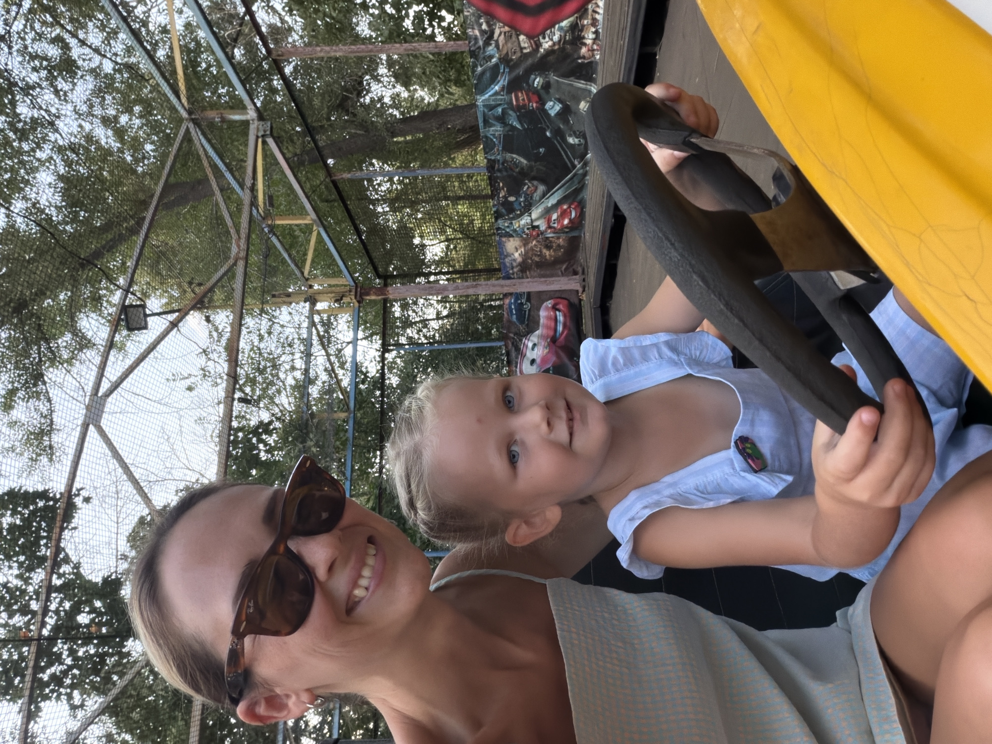 The image depicts a woman and a young child on an amusement park ride, likely a carousel or similar attraction. The woman, on the left, is wearing sunglasses and a light-colored dress. She is smiling broadly at the camera. The child, on the right, is holding a steering wheel and looking forward with a happy expression. The ride's backdrop features images of animated cars, possibly from the "Cars" franchise. The environment suggests an outdoor setting, with trees and a metal structure overhead, typical of an amusement park. The presence of specific amusement park rides and the overall atmosphere suggest a location like the amusement park at the City Park of Culture and Leisure in Kamyshin, Russia, a common venue for family entertainment.