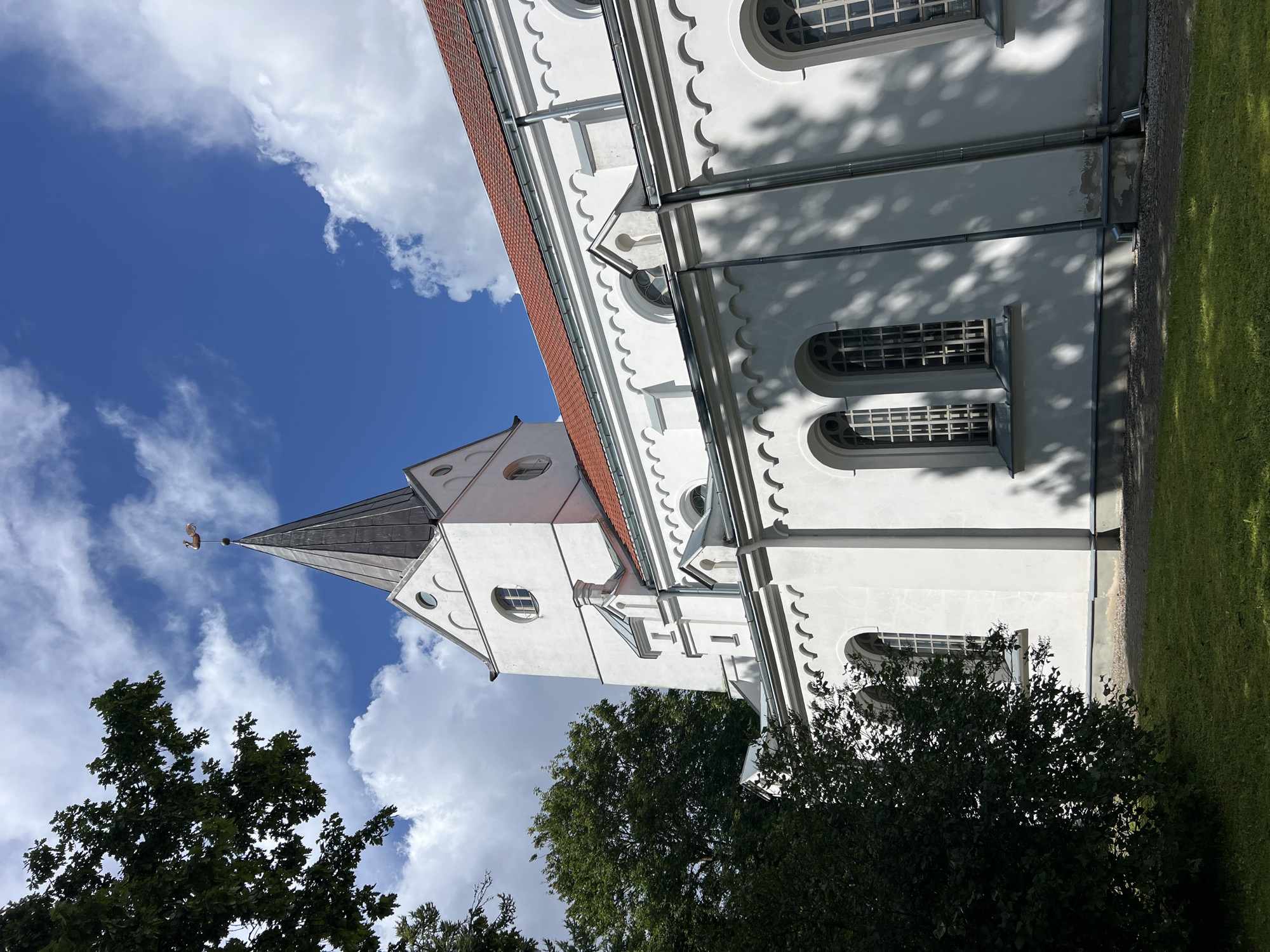 The image shows a white church with a red tiled roof and a dark spire topped with a weather vane depicting a swan. The church has arched windows with intricate leaded glass and decorative cornices. Trees with green foliage frame the left side of the building. The sky is blue with scattered white clouds. The church is located in the city of Saldus, Latvia. The ground in front of the church is covered with green grass and gravel. The lighting suggests it is daytime with sunlight casting shadows from the trees onto the church building. There are no people or animals visible in the image.