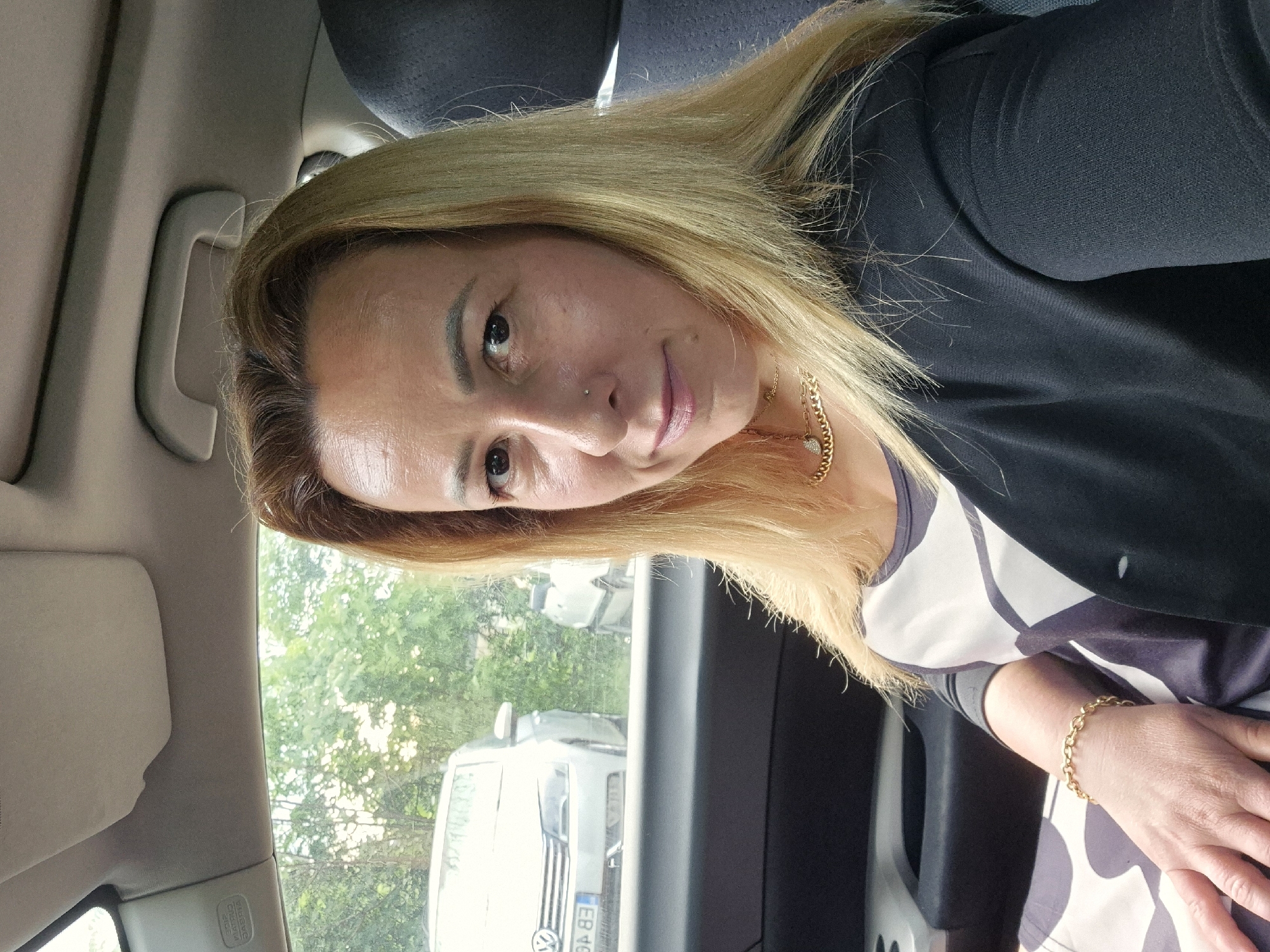 The image, rotated 90 degrees counter-clockwise, features a woman in what appears to be the front passenger seat of a car. She has long, straight blonde hair with visible darker roots and a clear complexion, looking directly at the camera with a neutral expression. A small stud nose piercing is visible on her right nostril.
She is wearing a dark, possibly black, open jacket over a short-sleeved top with a white and dark grey geometric pattern. Accessories include a gold chain necklace with a small heart-shaped charm and a gold link bracelet on her right wrist.
The car's interior is visible around her, including a light-colored grab handle above the window, a dark headrest, and part of a sun visor. Through the window, a bright outdoor scene is visible with green trees and another white vehicle. The white vehicle has a prominent Volkswagen (VW) logo on its grille and a partially visible license plate showing "EB 46" followed by two obscured characters. The setting appears to be daytime with natural light. A label reading "SRS AIRBAG" is partially visible near the car's pillar.