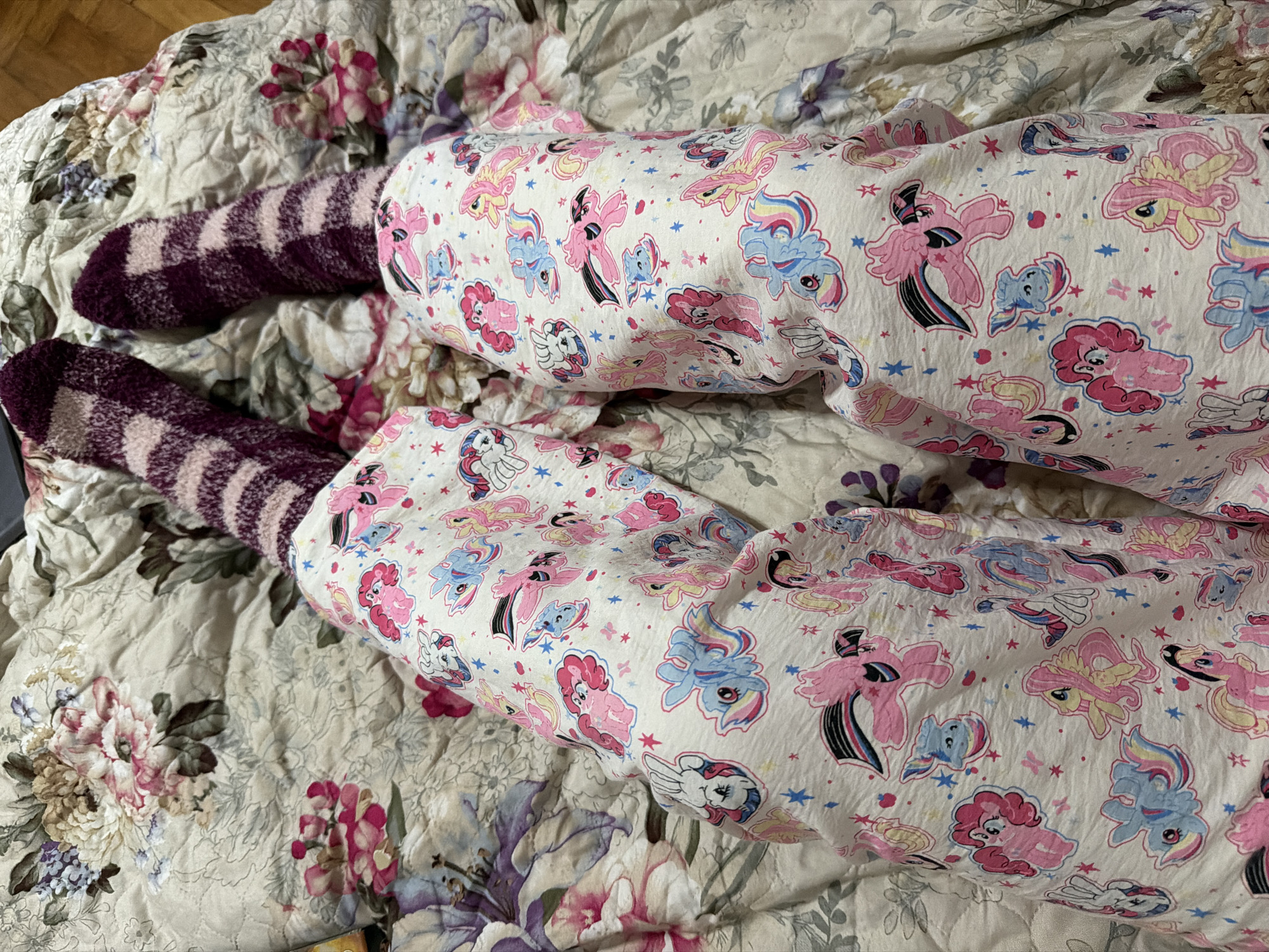 The image shows a person's legs covered in pink and white pajamas patterned with cartoon ponies, wearing purple and pink striped fuzzy socks, lying on a bed with a floral-patterned comforter. There is no information in the image that indicates the location in Moscow, Russia.