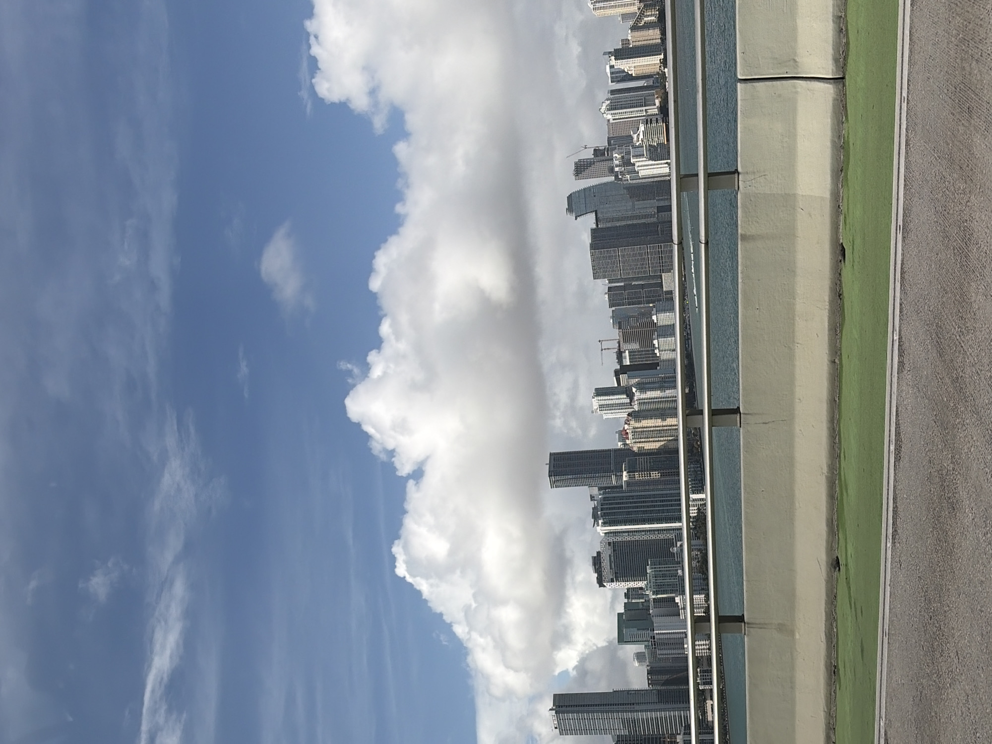 The image captures a view of the Miami skyline in the United States from an elevated perspective, potentially a bridge. To the left, a blue sky with scattered white clouds dominates the frame. Along the right edge, the concrete structure of the bridge is visible, with a green median strip and asphalt road. Beyond the bridge structure lies the cityscape of Miami, with numerous high-rise buildings and skyscrapers densely packed together. The buildings vary in height, color, and design, creating a varied architectural profile. The scene appears to be captured during daylight hours, with clear visibility.