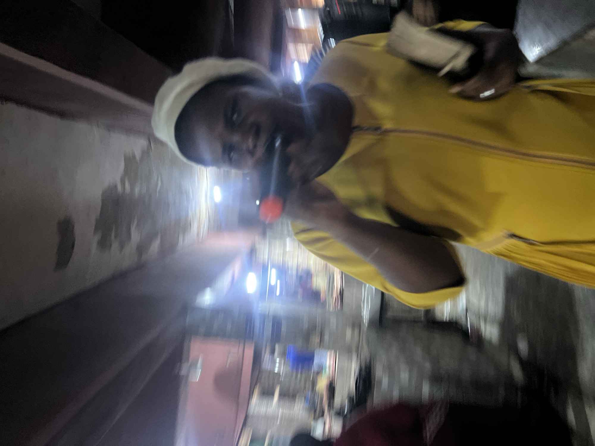 This image depicts a woman, likely in Nkpor, Nigeria, holding a microphone to her mouth, suggesting she is speaking or singing. She is wearing a light-colored head covering and a bright yellow garment with a zipper. In her right hand, she holds what appears to be a small book or pamphlet. The background is blurred, but suggests an indoor or covered outdoor market setting, with stalls and lighting visible. The overall lighting is dim, and the image has a motion blur, indicating movement during the capture.