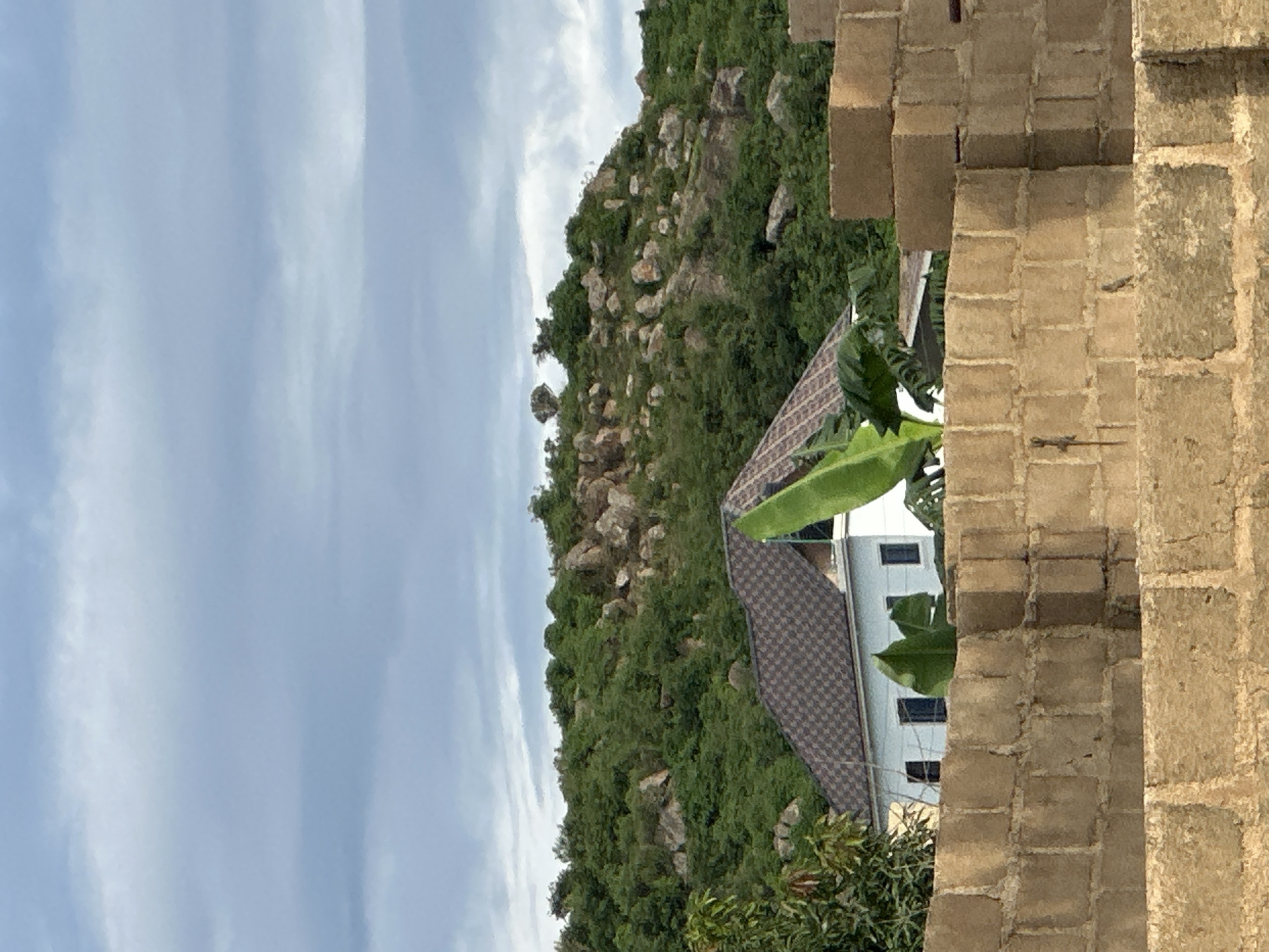 The image depicts a residential building situated in a hilly, vegetated area, likely on the outskirts of Jos, Nigeria. In the foreground, a wall constructed from concrete blocks forms a barrier, with the top section partially obscuring a two-story white house. The house features a dark, patterned roof and several rectangular windows. Lush green vegetation, including banana leaves, surrounds the house, with a glimpse of trees to the left. Behind the building, a rocky hill covered in dense green trees and scattered boulders rises against a cloudy sky. The overall impression is one of a semi-rural or suburban setting with a natural, rugged landscape.