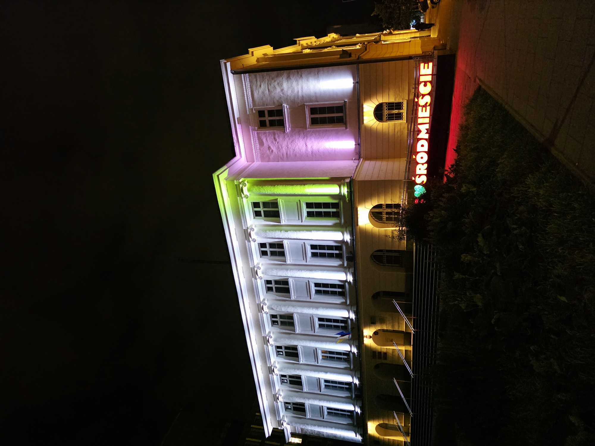 A night-time, low-angle shot depicts a large, well-lit building, likely a municipal or administrative structure, in Warsaw, Poland. The building is illuminated with a combination of white, green, and pink lights, highlighting its classical architectural features, including columns and arched entrances. A prominent neon sign reading "ŚRÓDMIEŚCIE" with a green heart next to the text is visible in the lower right portion of the image, illuminating the adjacent sidewalk and grass. The sign indicates the location is in the Śródmieście district of Warsaw. A lone figure, silhouetted against the light, walks on the sidewalk in the distance to the right. The foreground is dominated by dark, unkempt vegetation. The sky is completely black, indicating it is nighttime.