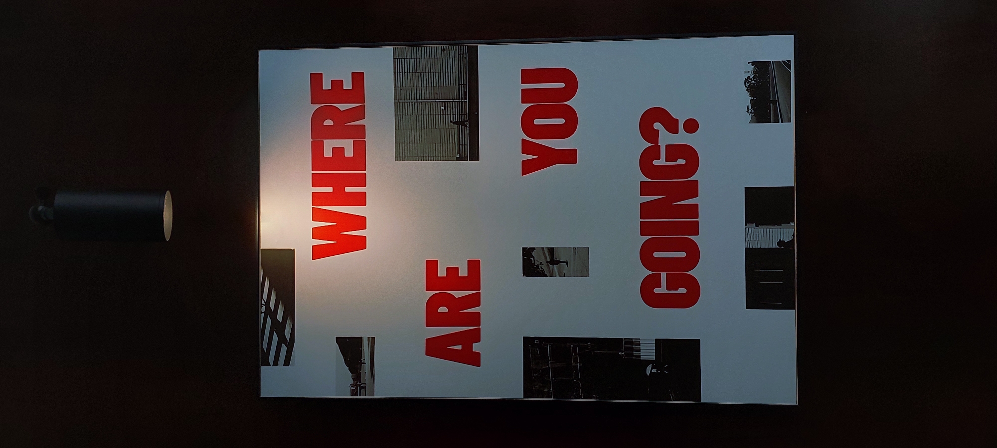 A framed artwork is displayed on a dark wall. The artwork features large, red, sans-serif text on a light-colored background, arranged in three lines. The text reads "WHERE ARE YOU GOING?". Interspersed among the text are several small, black and white photographs. One photo shows architectural elements of a building. Another depicts a single figure walking in front of a modern building. A third photo shows a person under an umbrella. Other photos show silhouetted figures in doorways and a street scene. A dark, cylindrical light fixture hangs from the ceiling directly above the artwork. The environment appears to be indoors, possibly a gallery or public space. There is no specific indication of the location being Pagedangan, Indonesia.