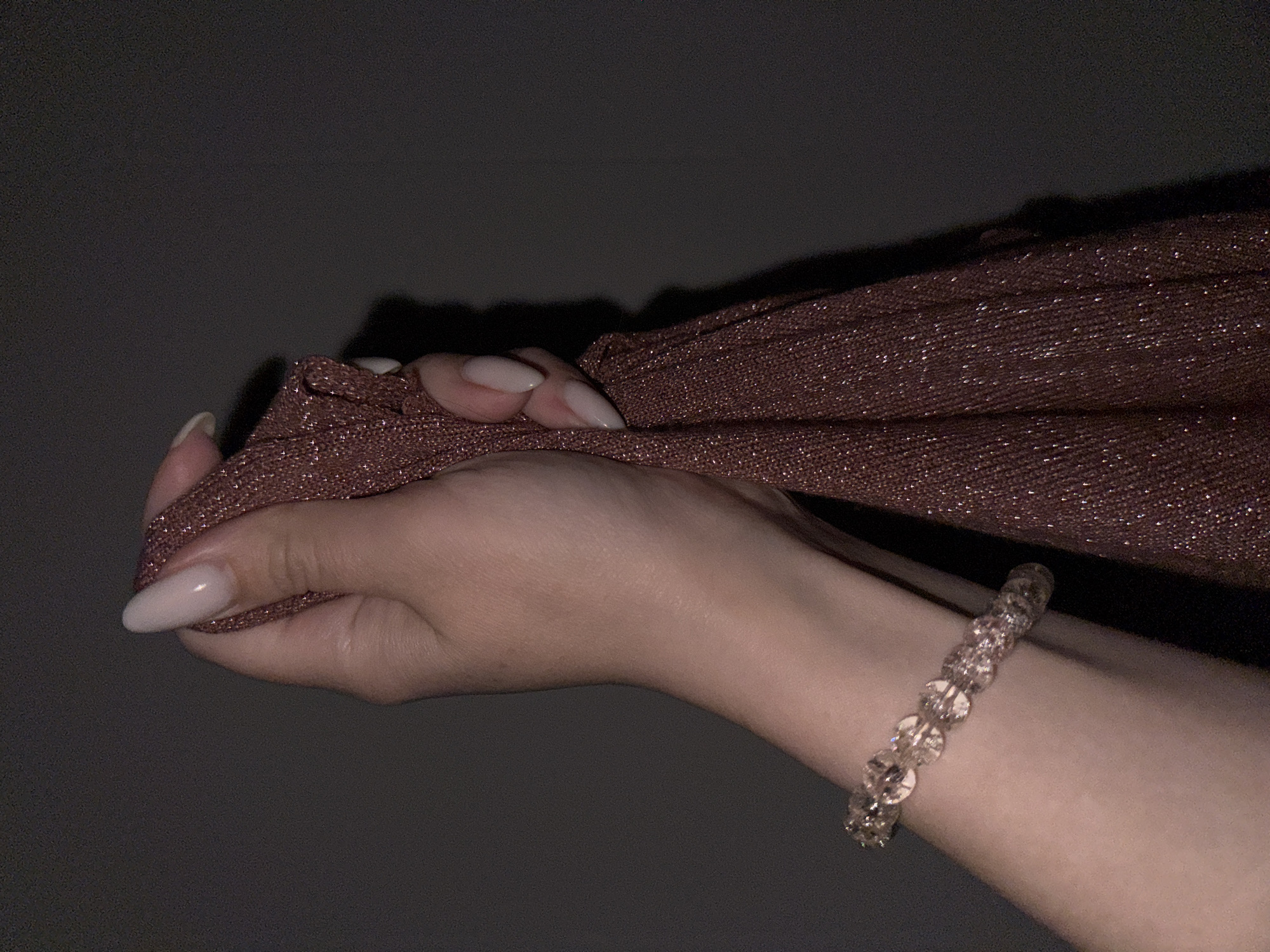 A close-up shot shows a person's left hand holding a shimmering brown fabric. The hand has long, almond-shaped nails with a light pink polish. A bracelet made of clear, faceted beads is worn on the wrist. The background is dark and indistinct. There is no evidence to suggest that the image was taken in Chaikovsky, Russia.