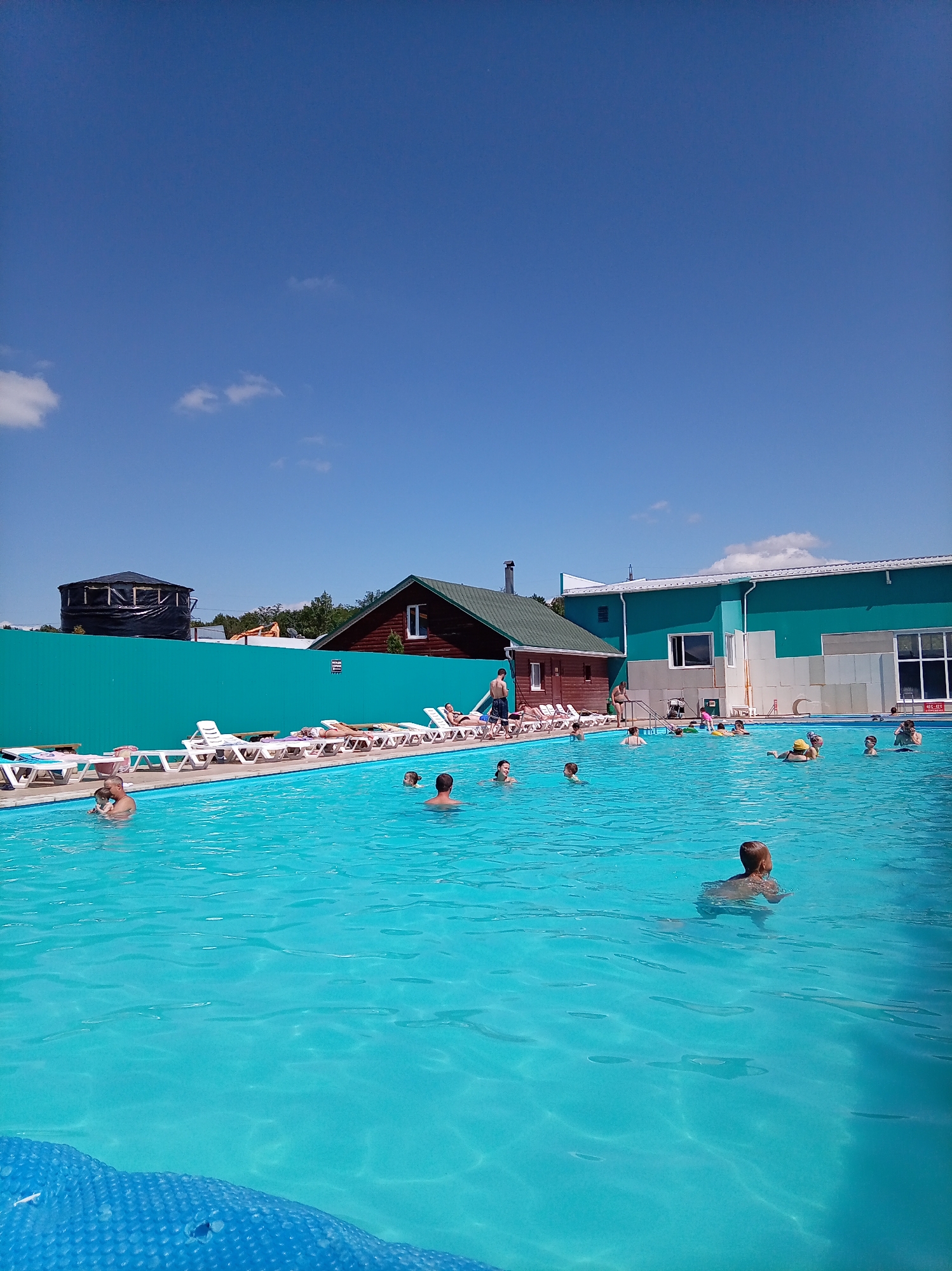 This is an outdoor daytime photograph taken at a swimming pool facility. The pool is filled with bright turquoise water, and several people are visible in and around it. In the water, there are individuals swimming, with some appearing to be adults and children. Along the edge of the pool, white lounge chairs are arranged, with some occupied by sunbathers. The background features a teal fence, a wooden building with a green roof, and a larger building with a teal and gray facade. There are also trees visible beyond the fence. A black, cylindrical structure covered with tarpaulin is also present in the distance. The sky is a clear, vibrant blue with a few scattered white clouds. The scene depicts a recreational activity in a sunny, outdoor environment. Based on the city of Кавказский, Russia, this could be a public or private pool complex offering leisure services.
