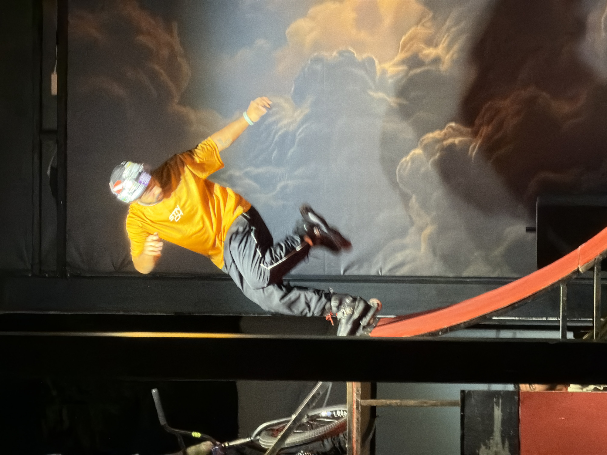 An athlete is performing a stunt on a ramp. They are wearing a yellow t-shirt, black pants, and rollerblades. The athlete is in mid-air, with their body contorted as if jumping. The background is a painted mural of clouds. The scene is indoors, likely in a performance venue. The city of Manavgat, Turkey, is not depicted in the image.