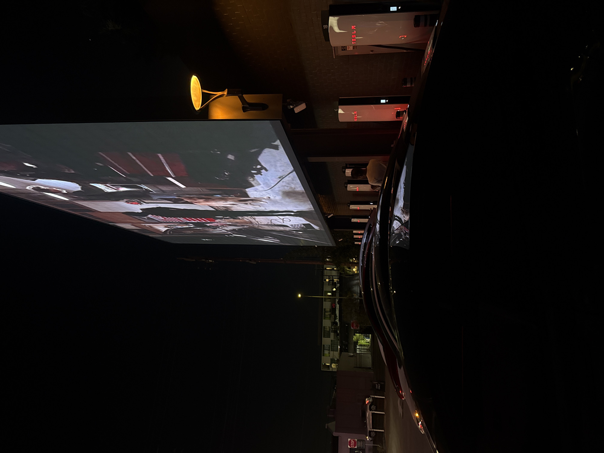 At night in West Hollywood, United States of America, a car is parked at a Tesla supercharging station. The station is adjacent to a large outdoor movie screen showing a film. A lamp is positioned near the charging stations. Tesla charging stations are visible, and street lights illuminate the surroundings. The scene is dominated by darkness.