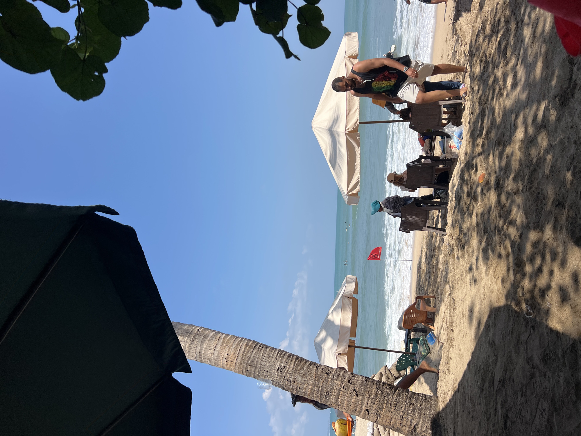 This image depicts a sunny day at Kuta Beach in Bali, Indonesia. The foreground is dominated by the sandy beach, with shadows from palm trees and umbrellas cast across it. In the midground, several beach umbrellas are set up, providing shade for beachgoers. People are seated at tables under the umbrellas, some appearing to be dining or relaxing. A man in a black tank top and white shorts is walking away from the camera, near an umbrella. Further out, the ocean stretches to the horizon under a clear blue sky. Waves are breaking on the shore, and a red flag is planted in the sand near the water's edge, possibly indicating swimming conditions. A coconut drink is visible on a tray held by a person near a palm tree on the left side of the frame. The overall atmosphere is one of a relaxed beach day.